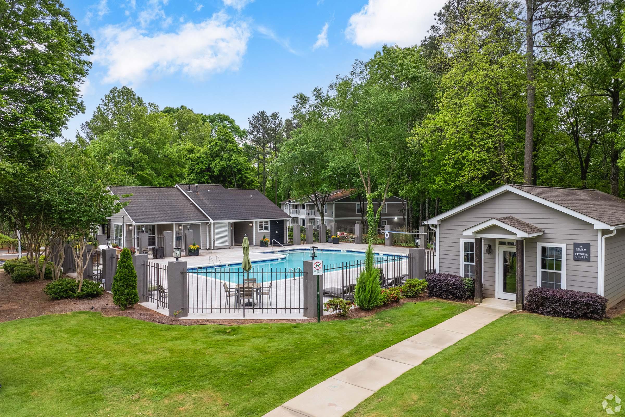 A serene outdoor scene featuring a swimming pool surrounded by lush greenery, a fence, and neatly landscaped grass. In the background, there are several buildings, including a cozy gray structure, and lounge chairs by the pool, creating a relaxing atmosphere.
