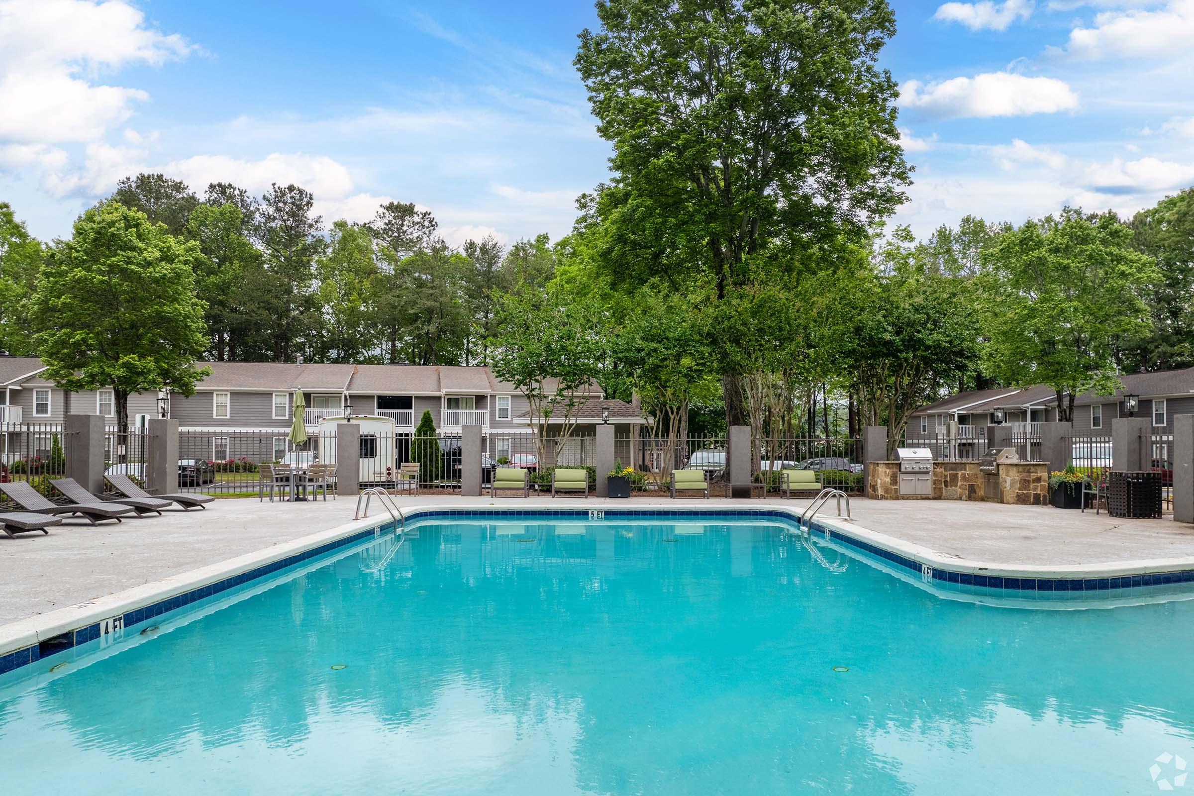 A clear swimming pool surrounded by lounge chairs and trees, with buildings in the background. The sky is blue with scattered clouds, creating a serene and inviting outdoor atmosphere.
