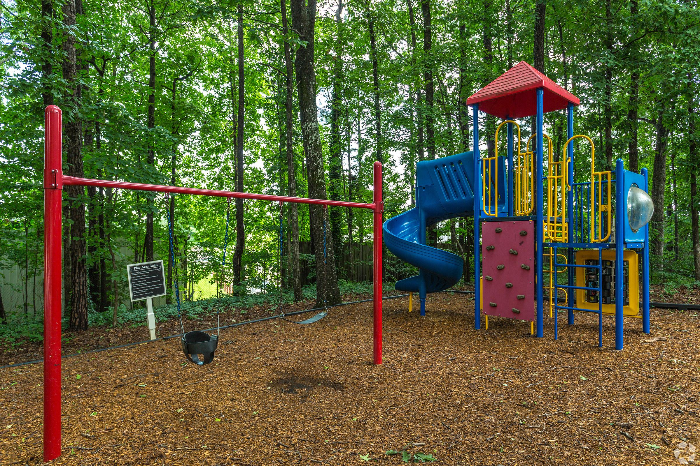 A colorful playground featuring a red swing set, a blue slide, climbing structures, and various interactive elements, surrounded by lush green trees and wood mulch on the ground. A sign is visible nearby, indicating park rules or information about the playground.