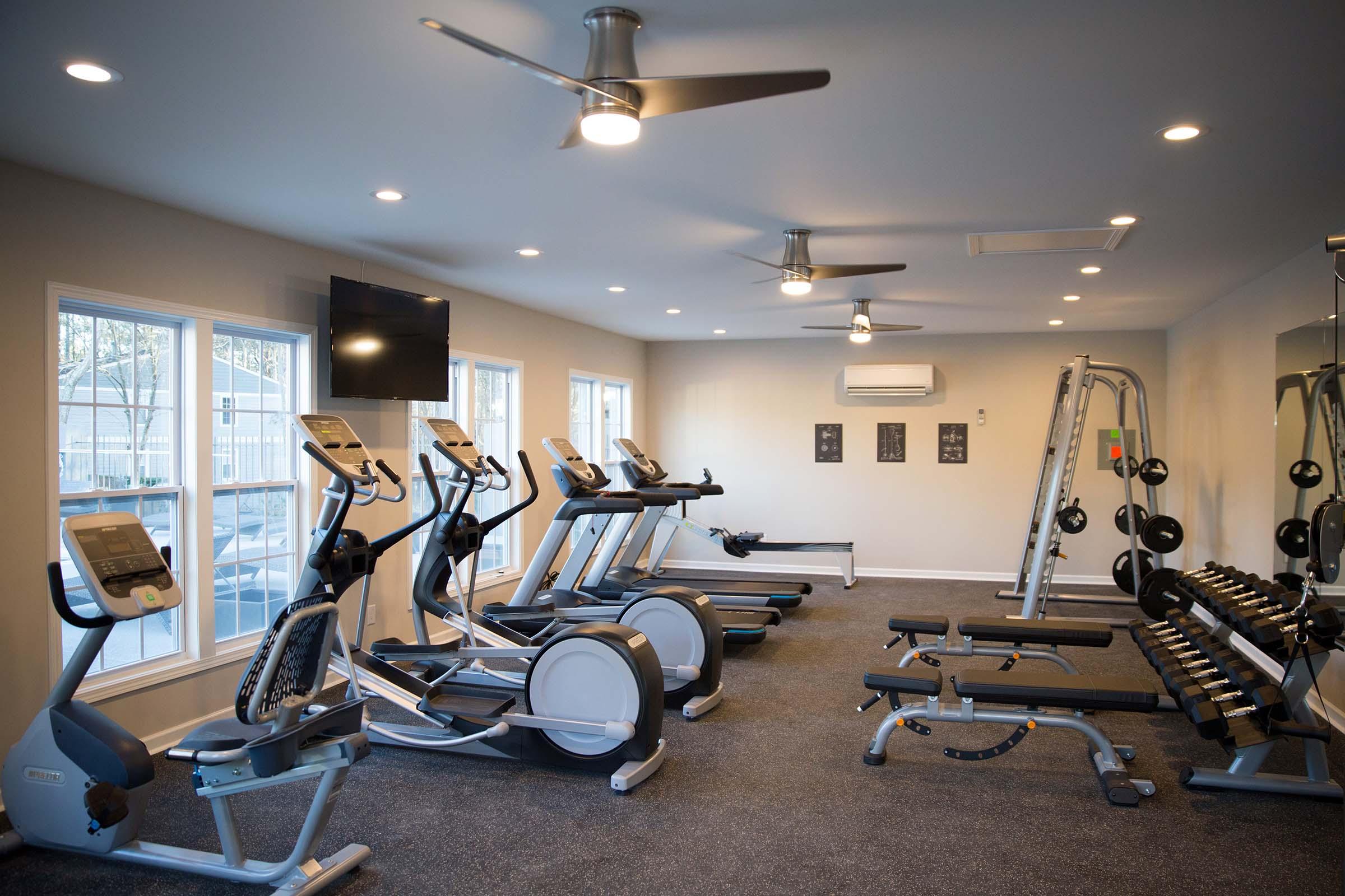 A modern gym interior featuring several cardio machines, including treadmills and ellipticals, along with a weightlifting area that has free weights and a weight rack. The room is well-lit with ceiling fans and large windows, creating a bright and inviting atmosphere for workouts.