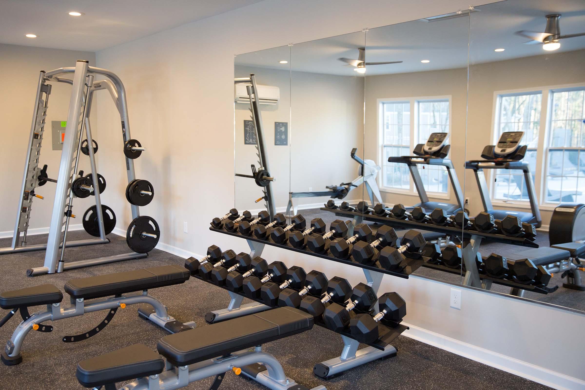 A fitness room featuring weightlifting equipment, including a weight bench, dumbbells arranged on a rack, and two cardio machines. The space is well-lit with large windows and mirrors, creating a modern and inviting workout environment.