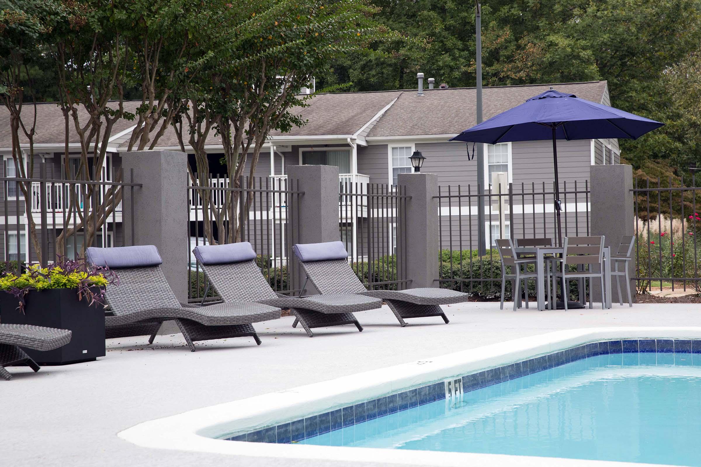 A clean outdoor swimming pool surrounded by lounge chairs and a small table with chairs. In the background, there are trees and a building with gray siding. An umbrella provides shade over the seating area, and a fence encloses the pool area for privacy.