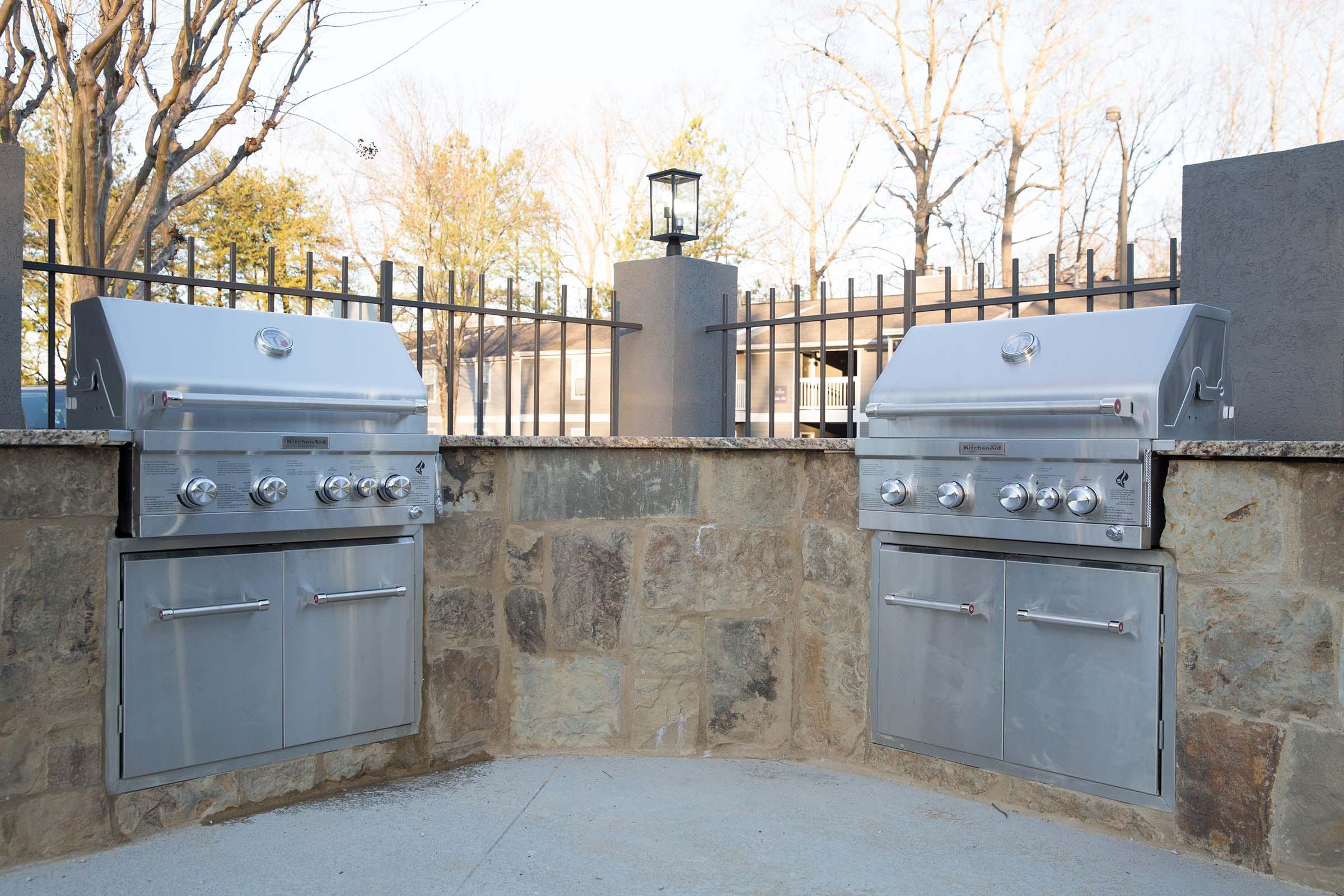 A well-designed outdoor kitchen featuring two stainless steel gas grills, set against a stone wall. The grills are mounted on a stone countertop with cabinets underneath. The area is surrounded by a wrought iron fence, and there is a lantern on a post nearby, with trees in the background.