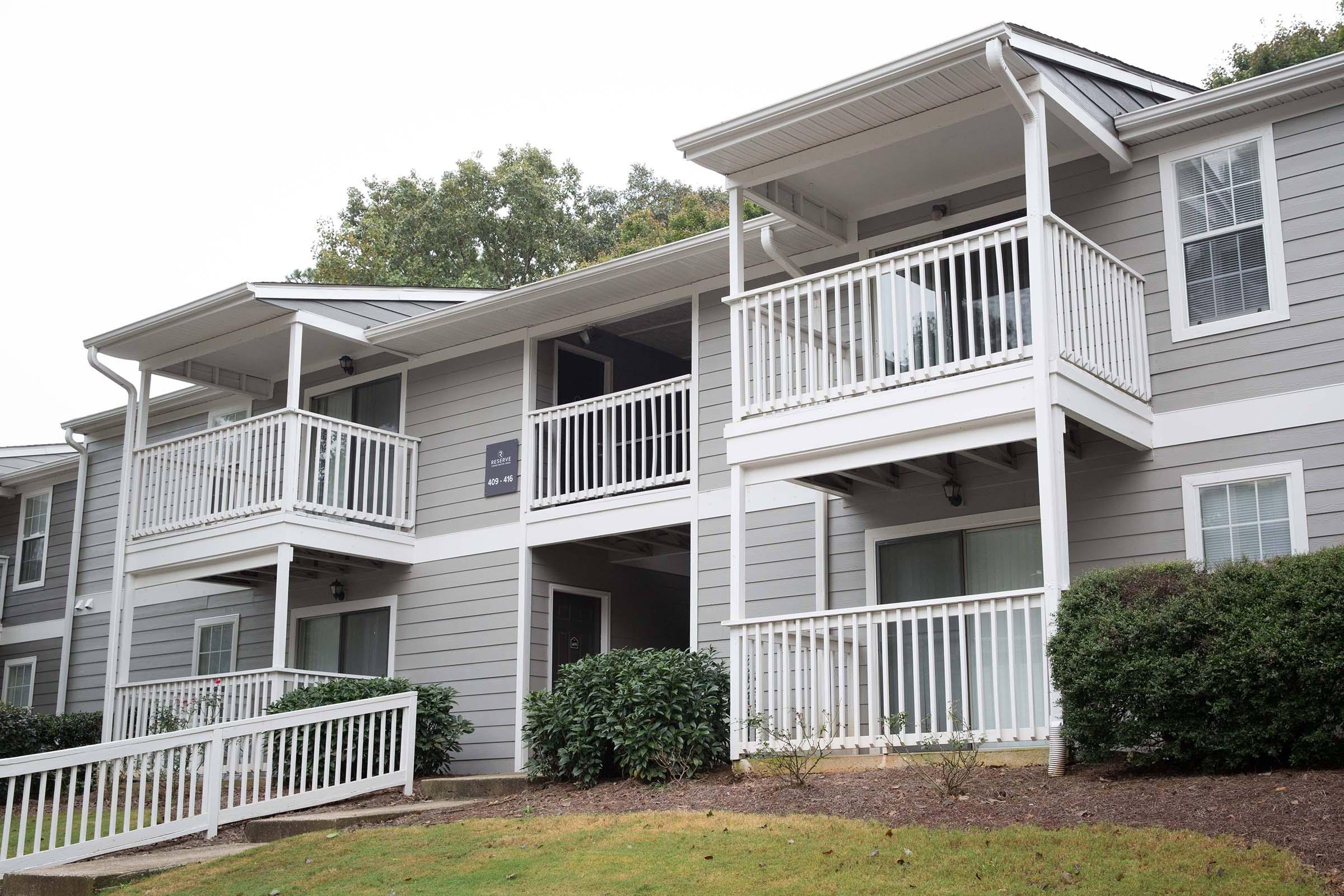 A multi-unit residential building with gray siding. The structure features two balconies with white railings, surrounded by green bushes and grass. The sky is overcast, indicating a cloudy day.
