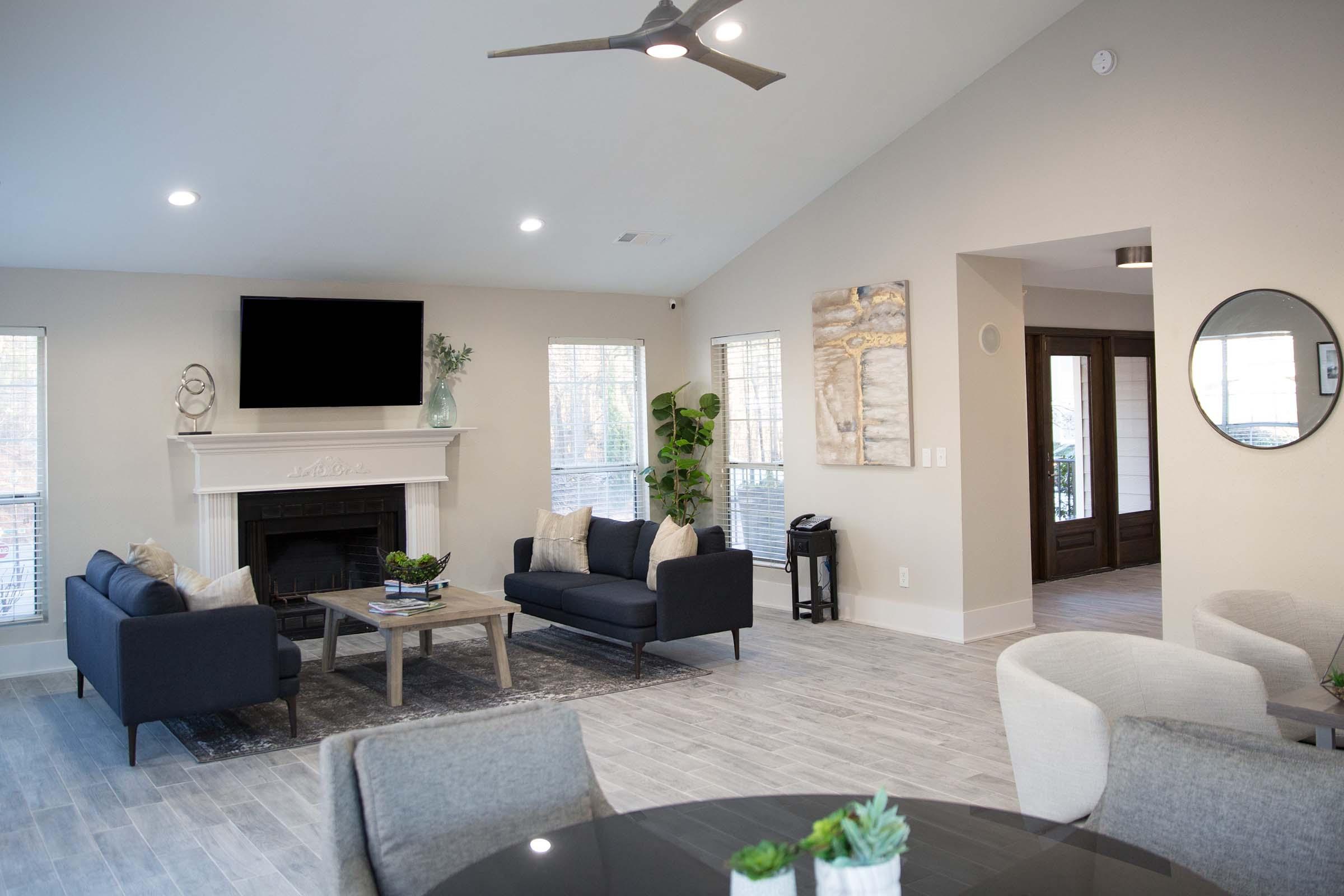 A modern living room featuring a neutral color palette with a large ceiling fan, comfortable couches, a coffee table, and decorative plants. A fireplace is present with a television mounted above it. Large windows allow natural light, and an adjacent dining area includes a round table and chairs.
