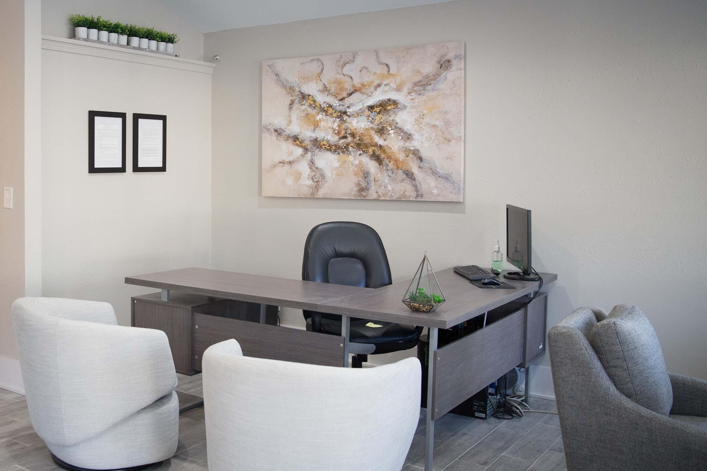 A modern office interior featuring a sleek gray desk with a black chair, a computer, and decorative items. The walls are light-colored, adorned with a large abstract painting and two framed documents. Plush white and gray chairs are arranged around the desk, providing a comfortable atmosphere.