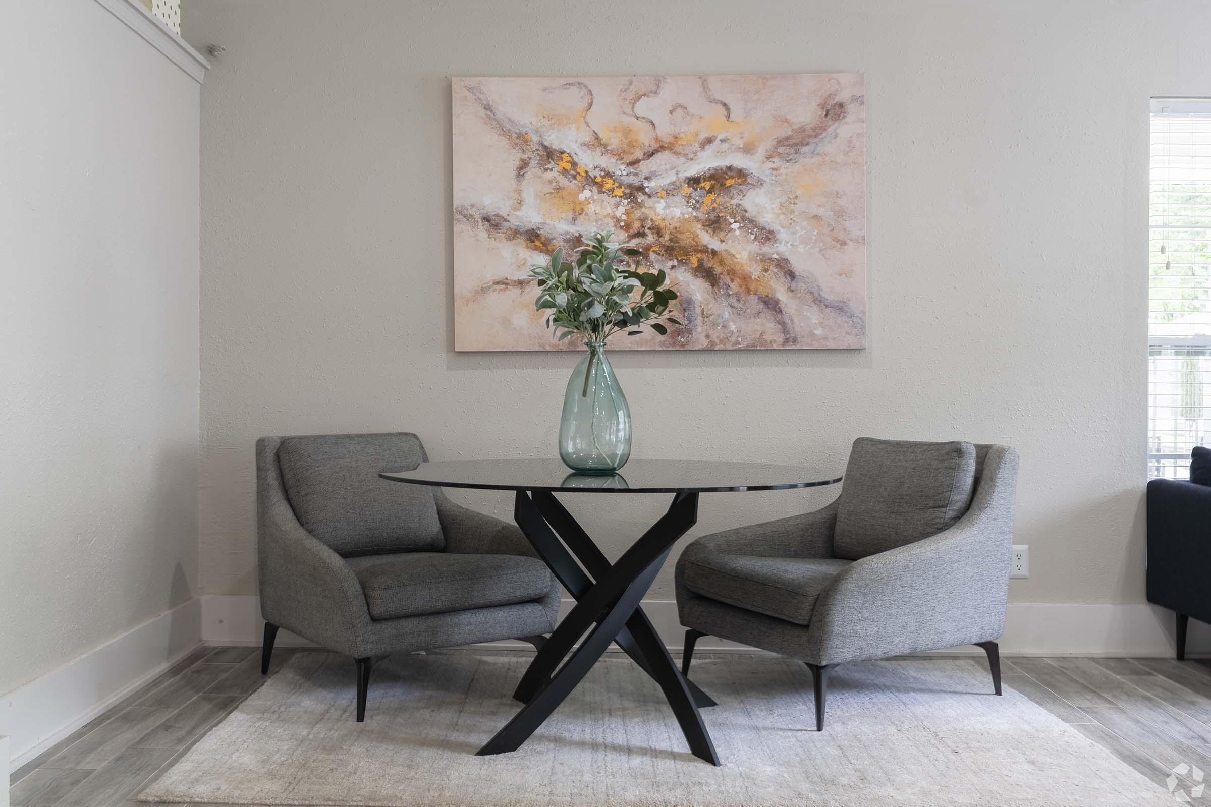 A modern living room setting featuring two gray armchairs with a round black coffee table in between. A decorative vase with greenery sits on the table. Behind them, there is an abstract painting in soft colors hanging on the wall and soft gray carpeting underfoot. Natural light filters in through a window.