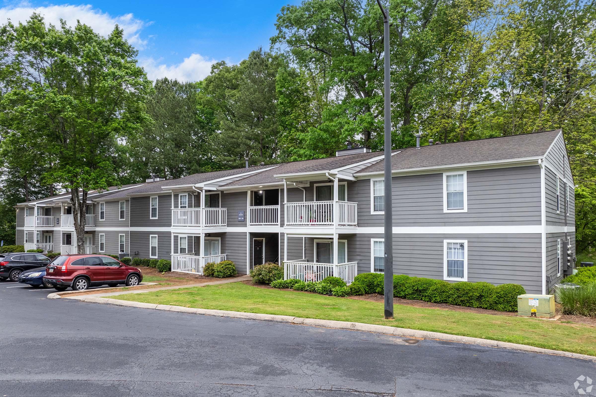 A multi-unit apartment building featuring grey siding and white balconies, surrounded by green trees and landscaped grounds. A parking area is visible in front, with several cars parked along a curved drive. The sky is clear with a few clouds.