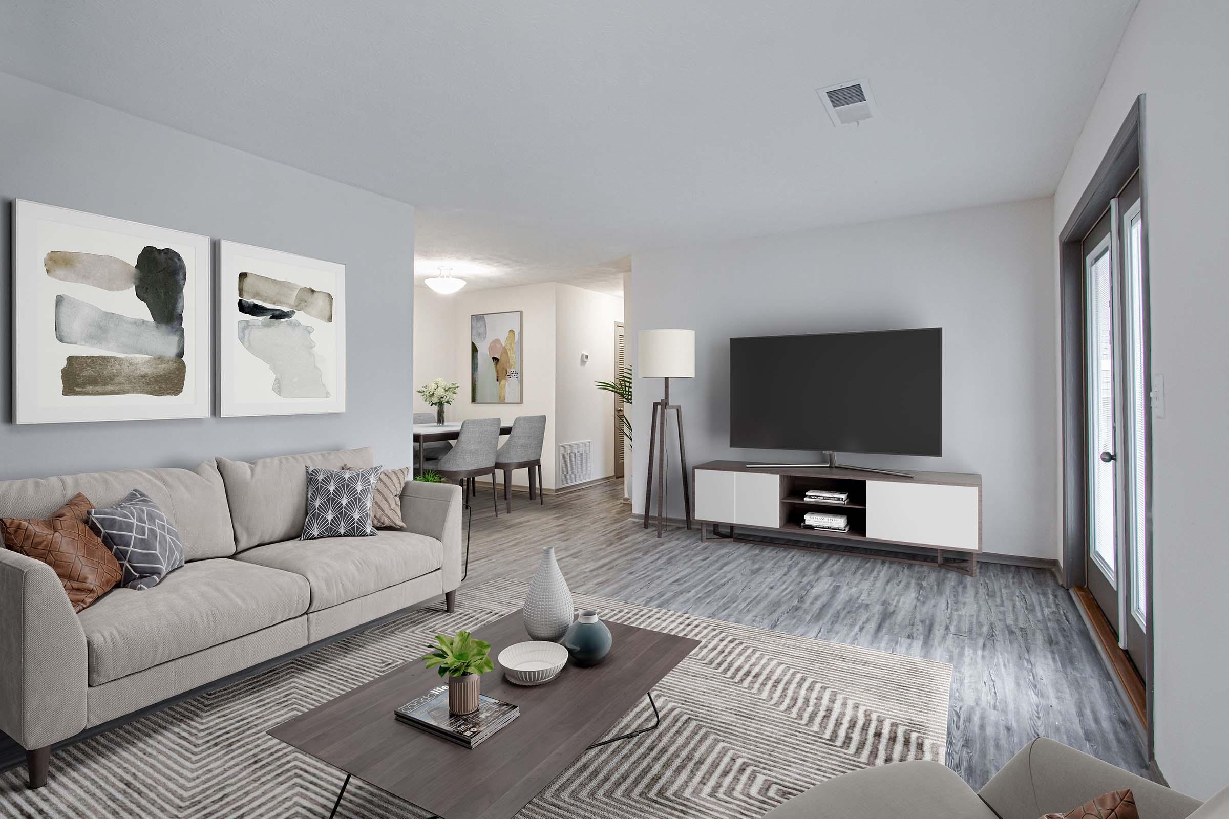 A modern living room featuring a beige sofa with decorative pillows, a coffee table with a plant and decorative items, and a large TV on a white cabinet. The walls are light gray, adorned with abstract artwork. In the background, a dining area with a table and chairs is visible, along with a door leading outside.