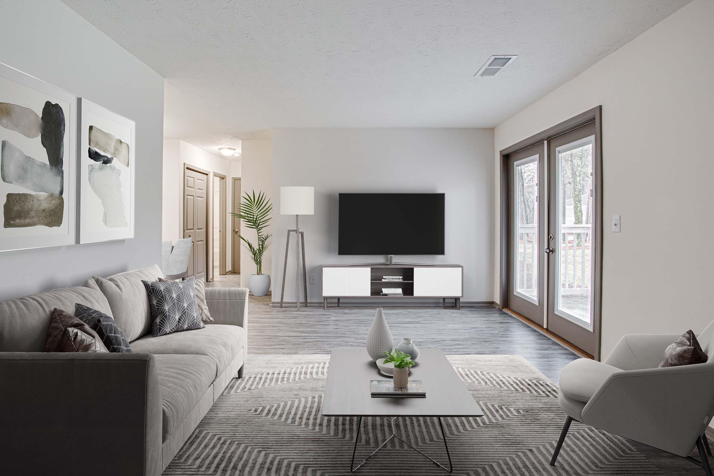 A modern living room featuring a light gray sofa with decorative pillows, a coffee table with plants, and a flat-screen TV on a white media console. There are light-colored walls, a floor lamp, and large windows with a view of greenery outside. The decor is minimalistic and contemporary.
