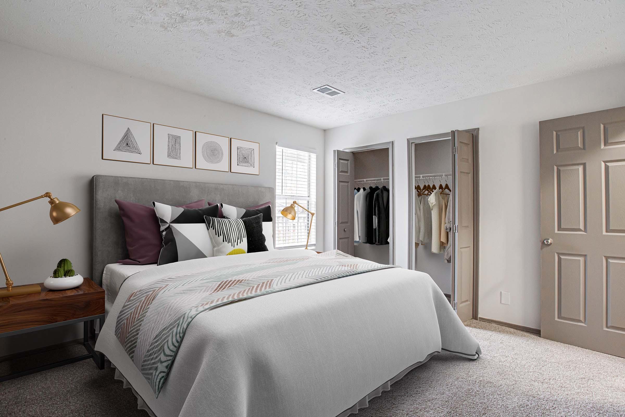A cozy bedroom featuring a gray upholstered bed with decorative pillows and a light gray blanket. The room has neutral walls, a window with blinds, and framed art prints above the bed. A wooden nightstand with a lamp sits beside the bed, while a closet with hanging clothes is visible in the background.