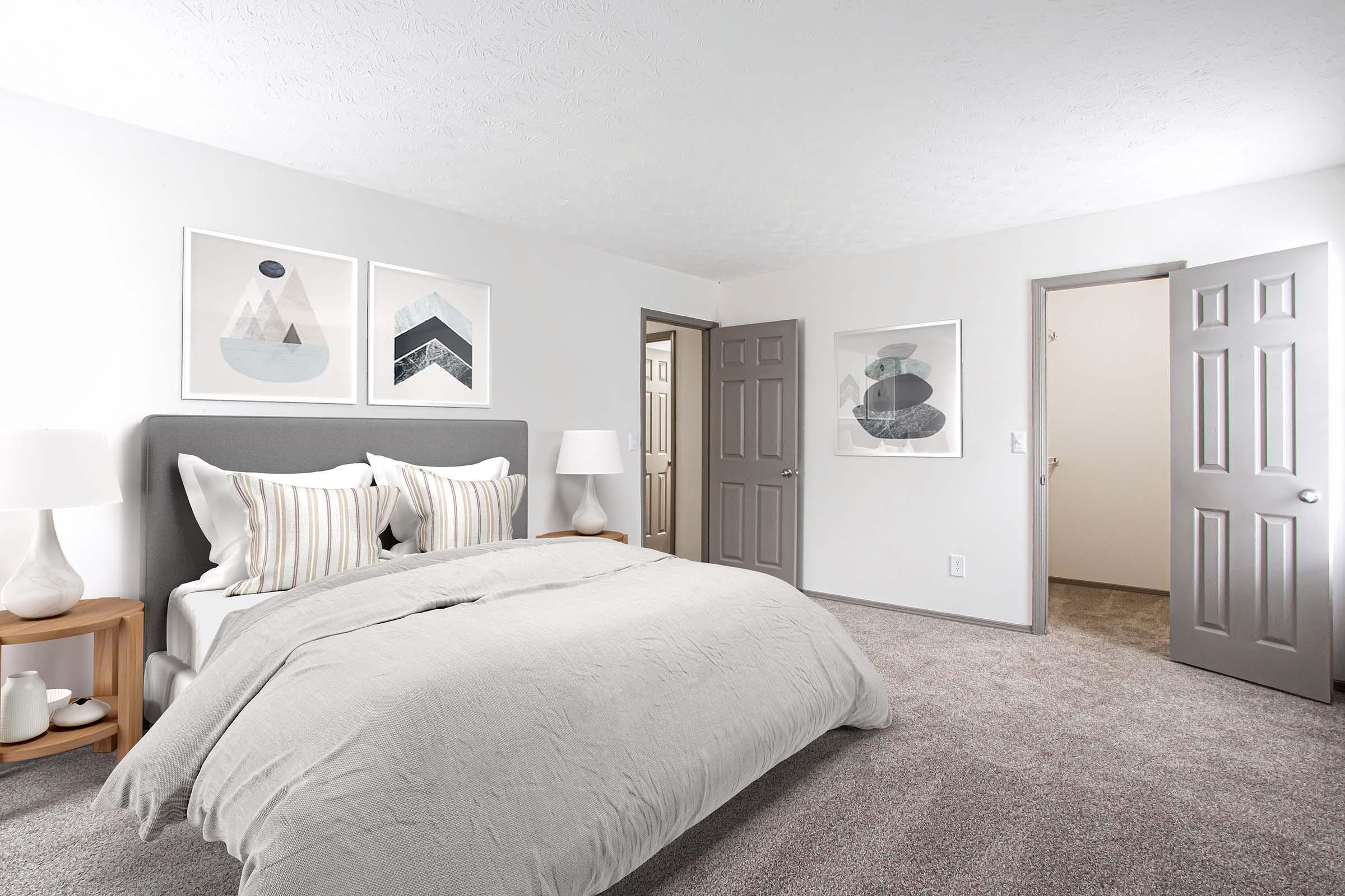 A neatly organized modern bedroom featuring a soft gray bed with striped pillows, light wood nightstands with lamps, and abstract wall art. The walls are painted light gray, and there are two closed doors in the background, one leading to another room. The carpet is plush and beige.