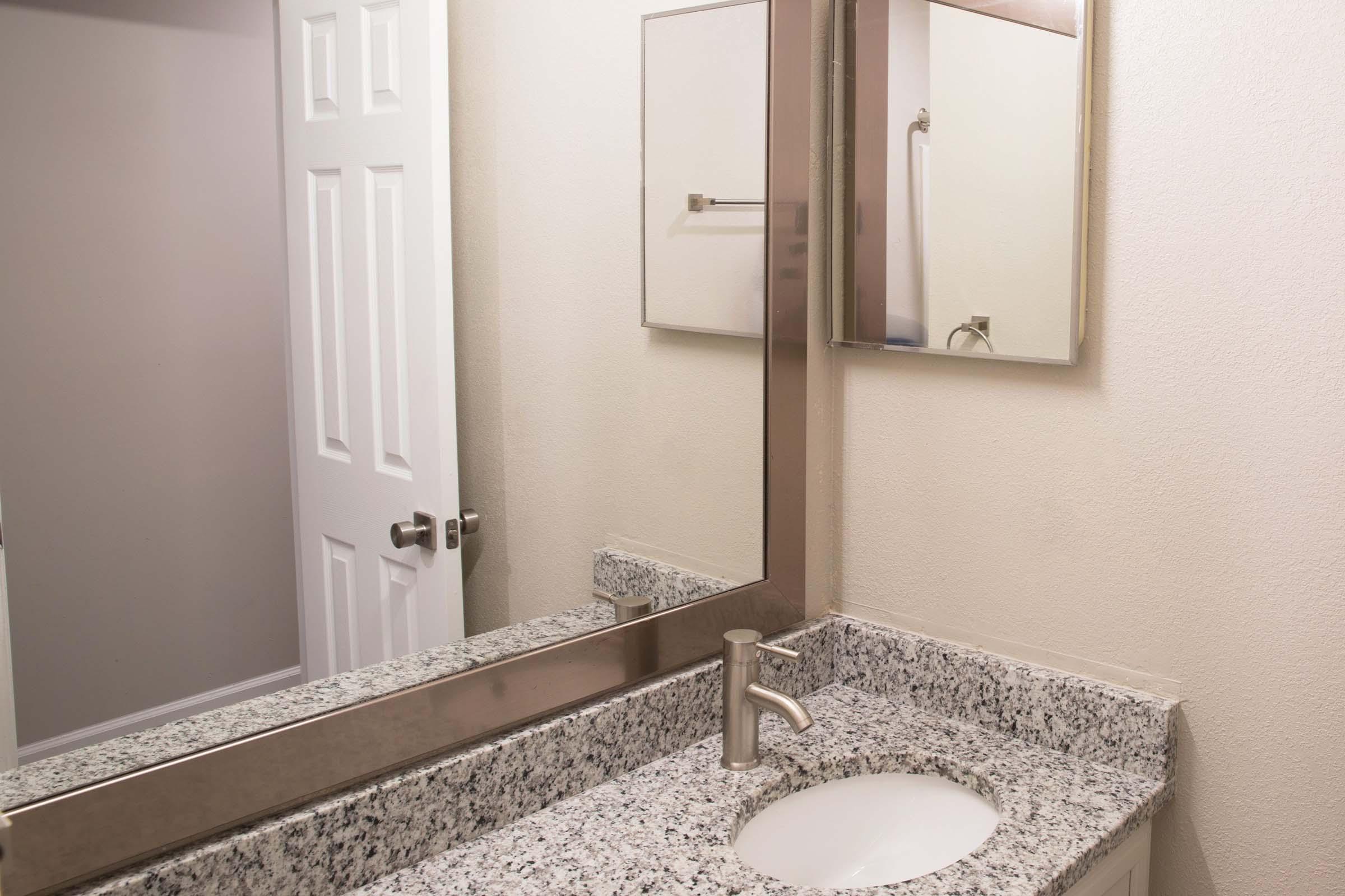 A clean bathroom with a granite countertop and a circular sink. A large mirror is mounted above the sink, reflecting the space. A door is visible in the background, and the walls are painted a light color, contributing to a bright and airy atmosphere.