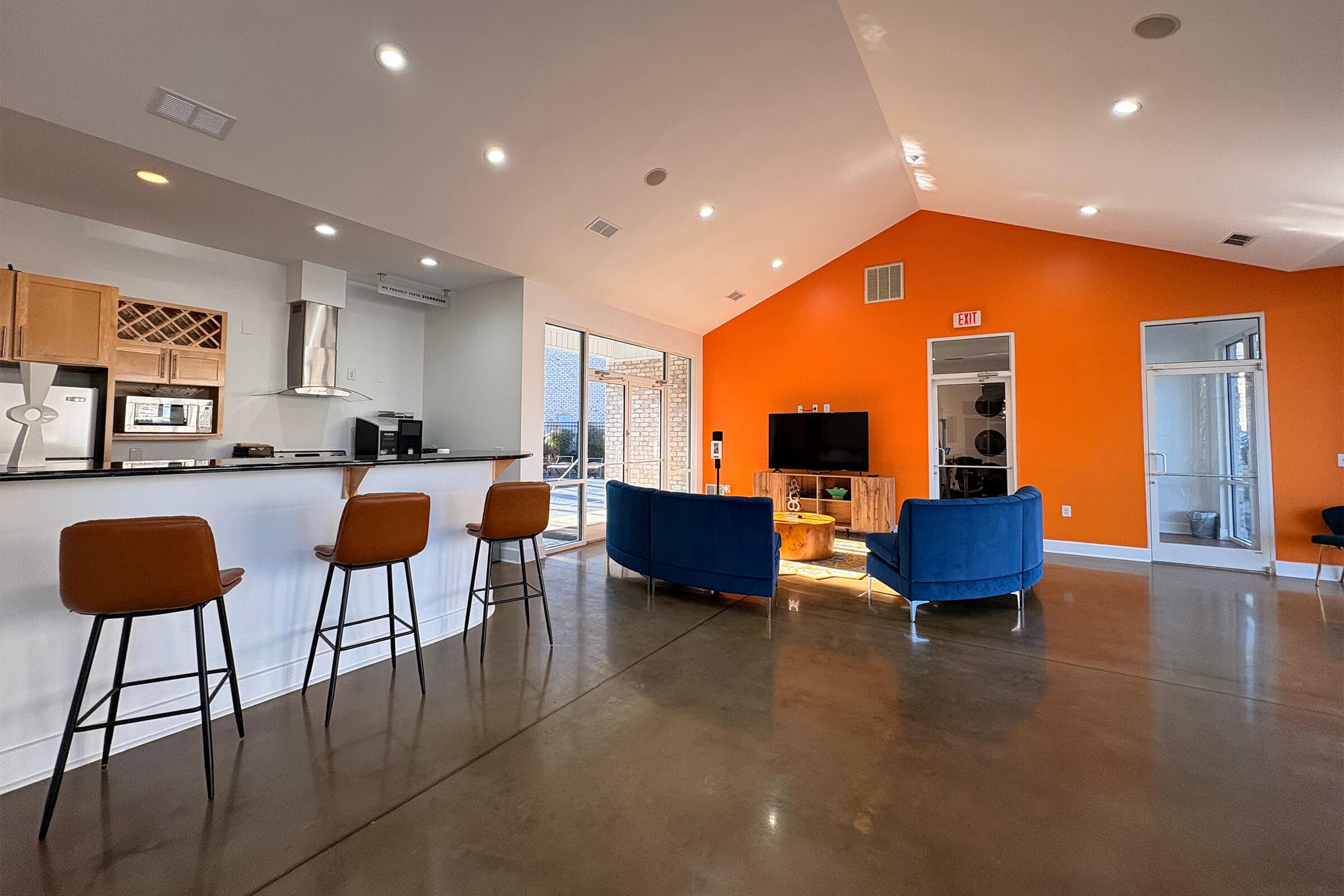 A modern living space featuring a kitchen bar with high stools, a cozy lounge area with blue chairs and a wooden coffee table, and an orange accent wall. Large windows allow natural light, and the polished concrete floor adds a contemporary touch.
