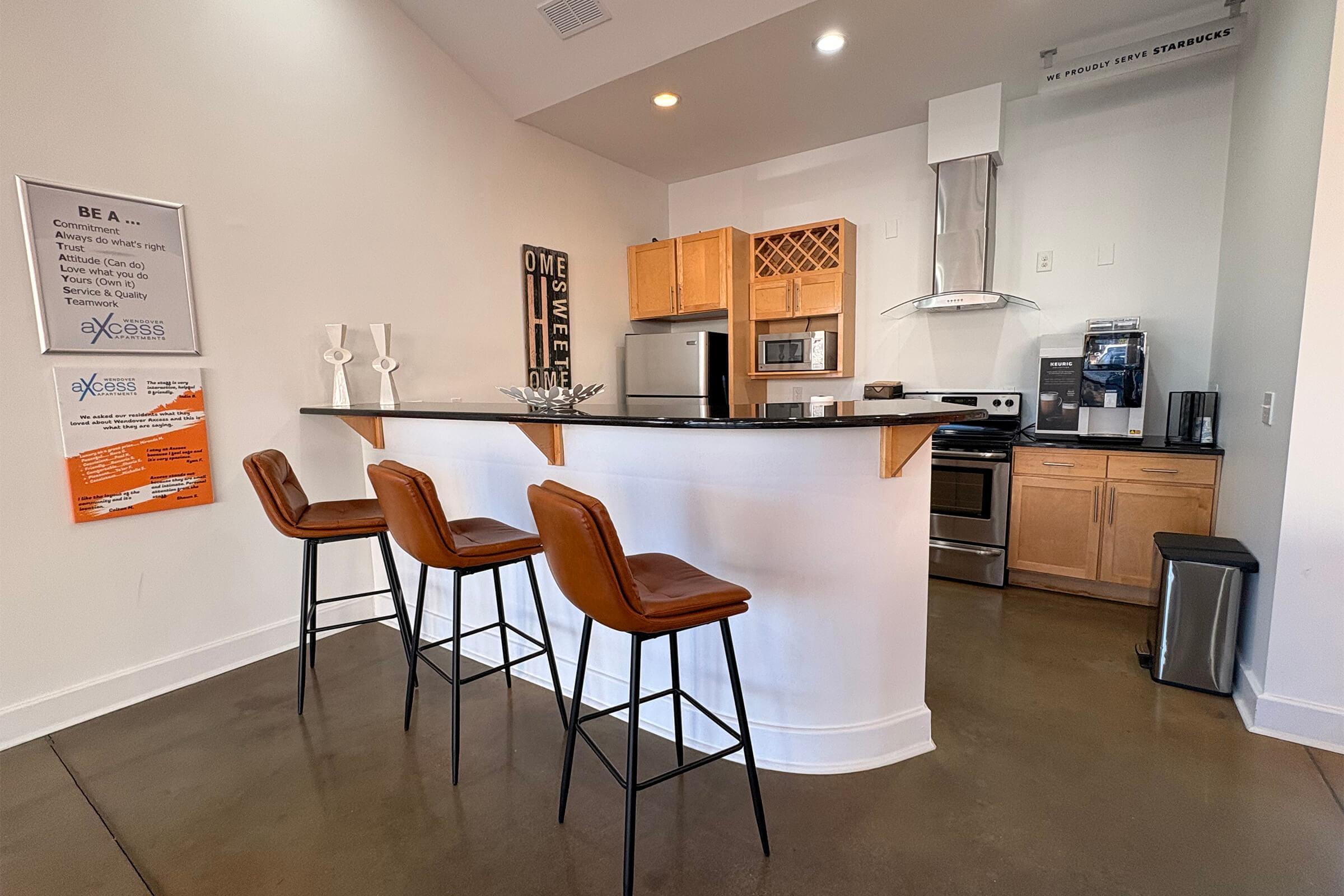 Modern kitchen with a central island, featuring three brown bar stools. Includes stainless steel appliances like a refrigerator, stove, and coffee machine. Light wood cabinetry and a black countertop. Wall art with the words "HOME SWEET HOME" and a poster on the side with information about services.