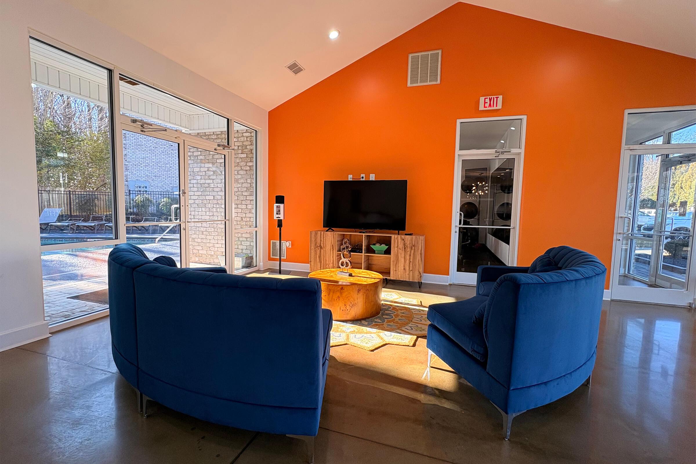 A bright, modern lounge area featuring two blue chairs facing a wooden coffee table, with a television mounted on the wall. The walls are partially orange, and large windows allow natural light to fill the space, revealing a view of an outdoor pool area.