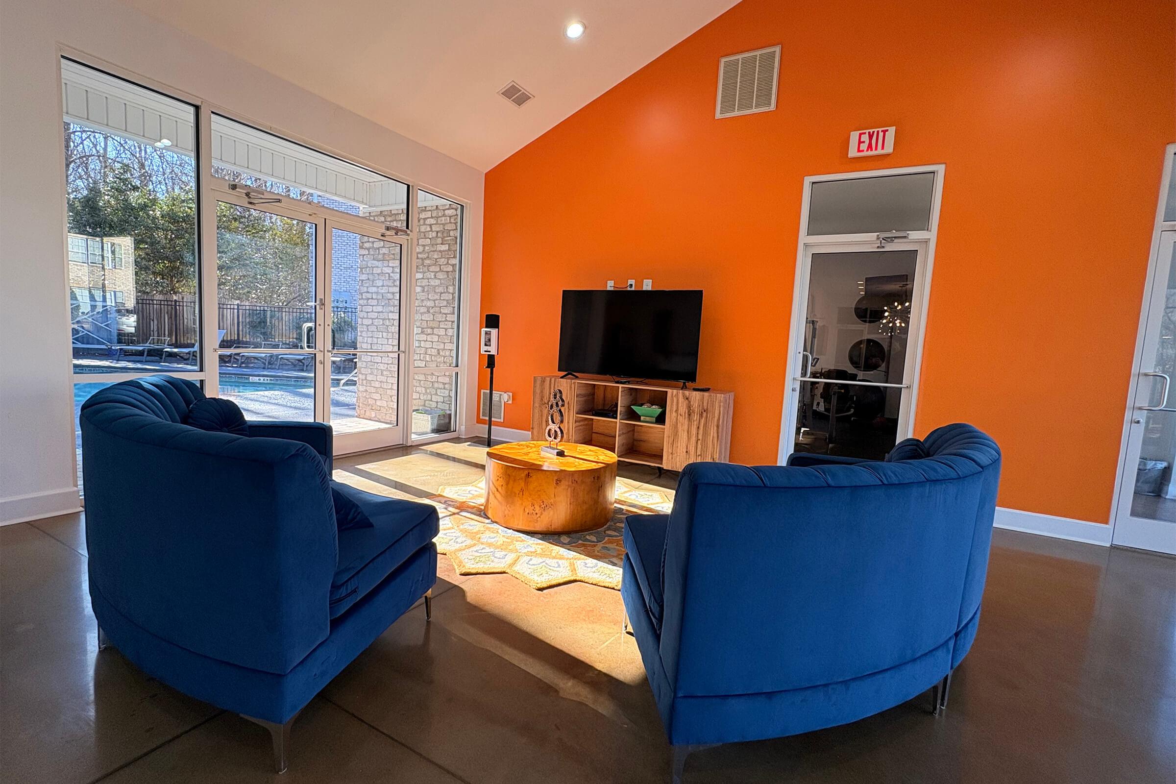 Brightly lit room featuring two blue chairs facing a wooden coffee table with a patterned rug underneath. A large TV is mounted on a wood cabinet against an orange accent wall. Large glass doors lead to an outdoor area, allowing natural light to fill the space.