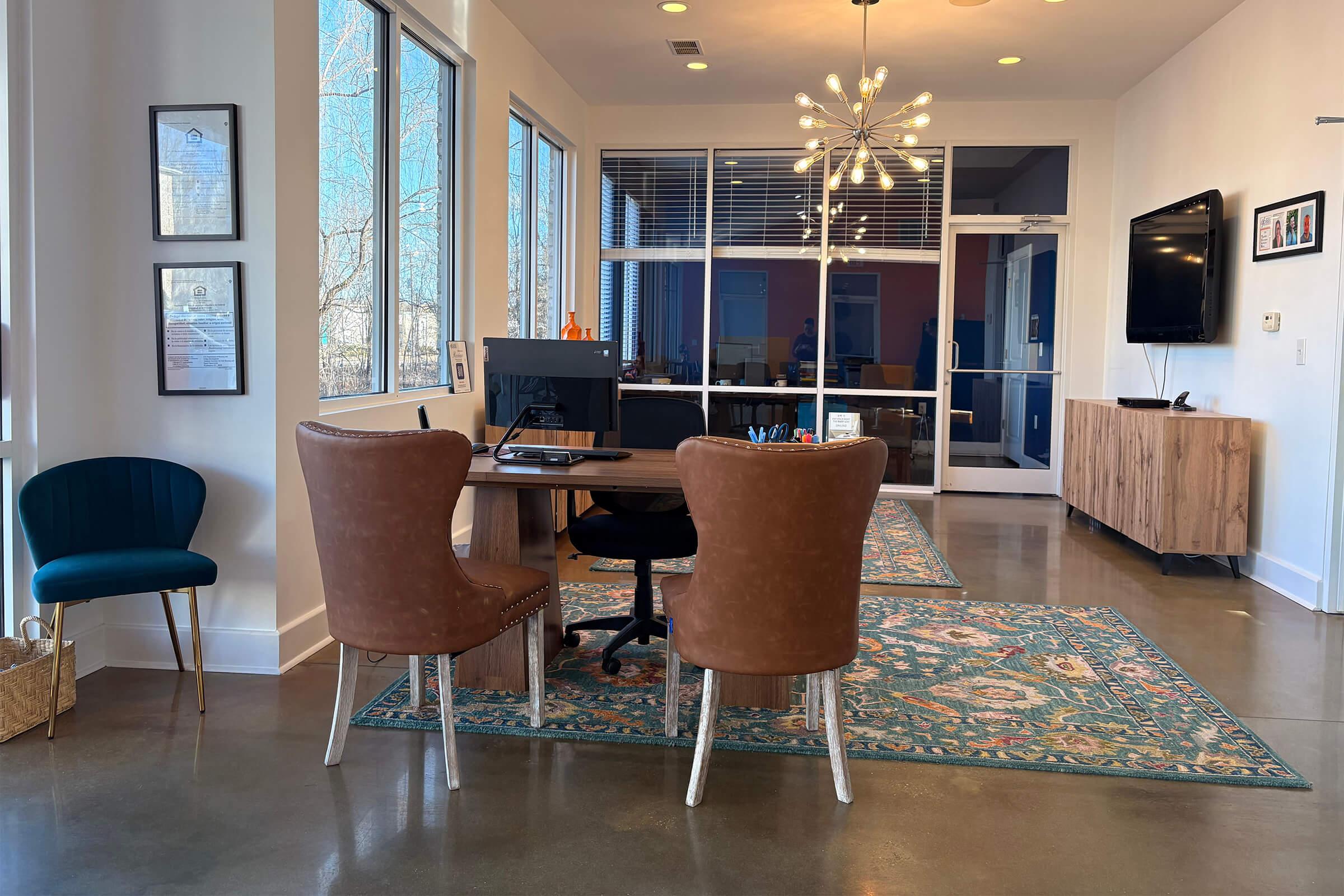A modern office interior featuring a wooden desk with a computer and stationery, two stylish brown armchairs, a teal accent chair, and a patterned rug. Large windows allow natural light to fill the space, and there’s a decorative light fixture overhead. A media console with a TV is also visible.