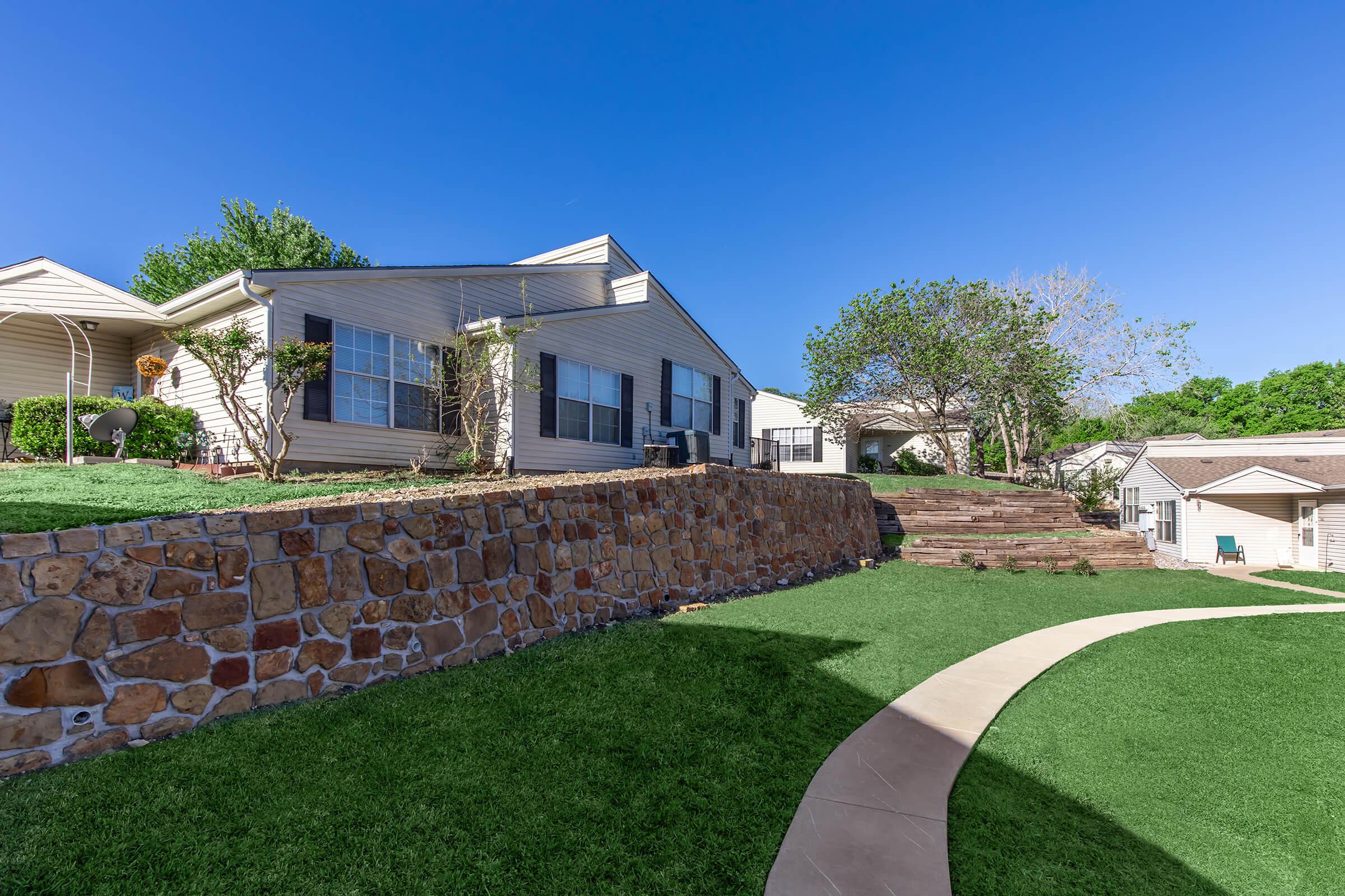 A residential area featuring a house with a stone wall in the foreground, a neatly landscaped lawn, and a curved pathway. The sky is clear blue, and there are trees and other buildings in the background, creating a peaceful suburban atmosphere.