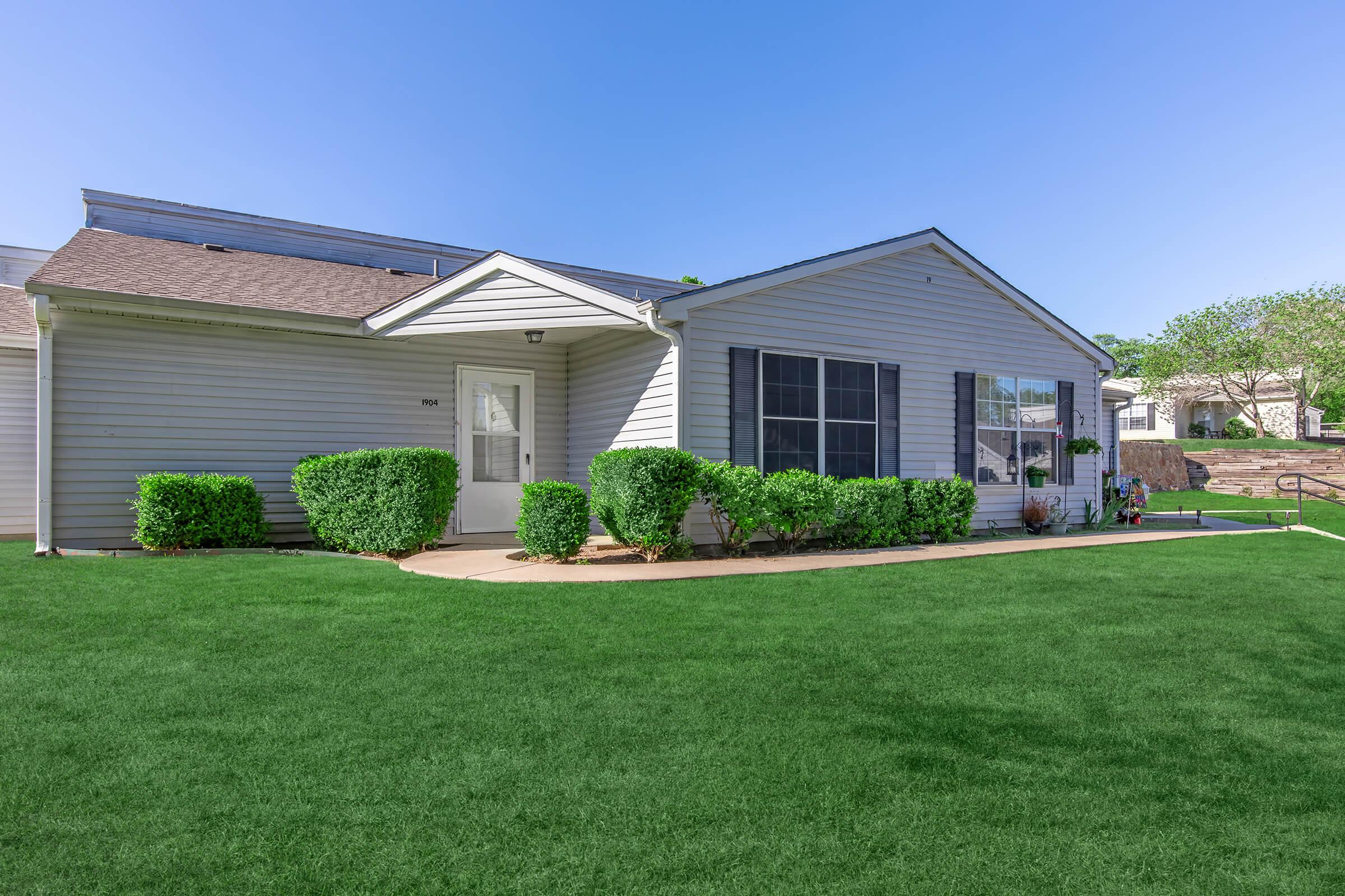 A well-maintained single-story house with gray siding and a brown roof. The front features neatly trimmed hedges and a vibrant green lawn. Large windows are adorned with black shutters, and a pathway leads to the entrance. In the background, other residential buildings and greenery are visible under a clear blue sky.