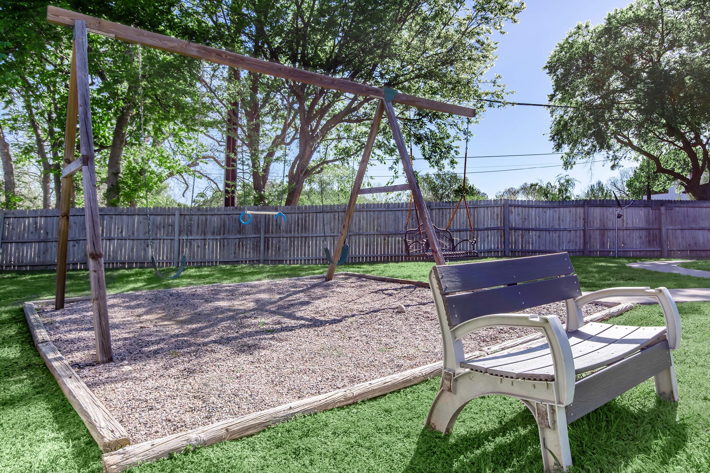 A sunny playground featuring a wooden swing set with two swings, surrounded by lush green trees and grass. In the foreground, there’s a wooden bench facing the swings, sitting on a gravel surface, with a wooden fence visible in the background.