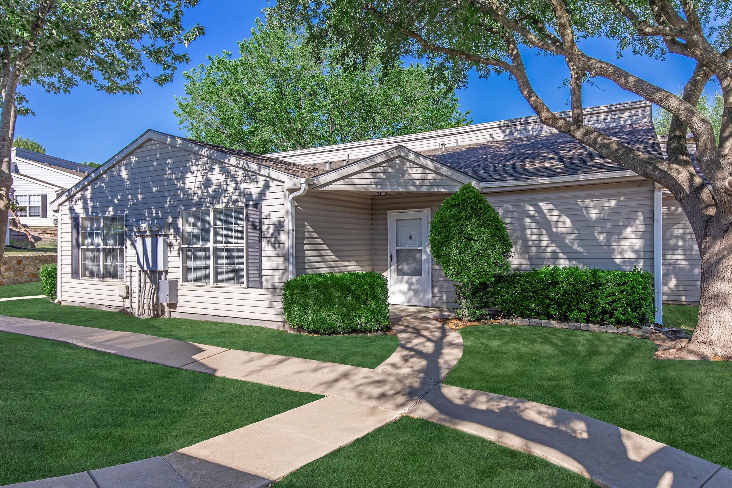 A light-colored single-story house with a white door and large windows, surrounded by green grass and shrubs. A concrete walkway leads to the entrance, and shade is provided by nearby trees. Bright blue sky complements the verdant scene.
