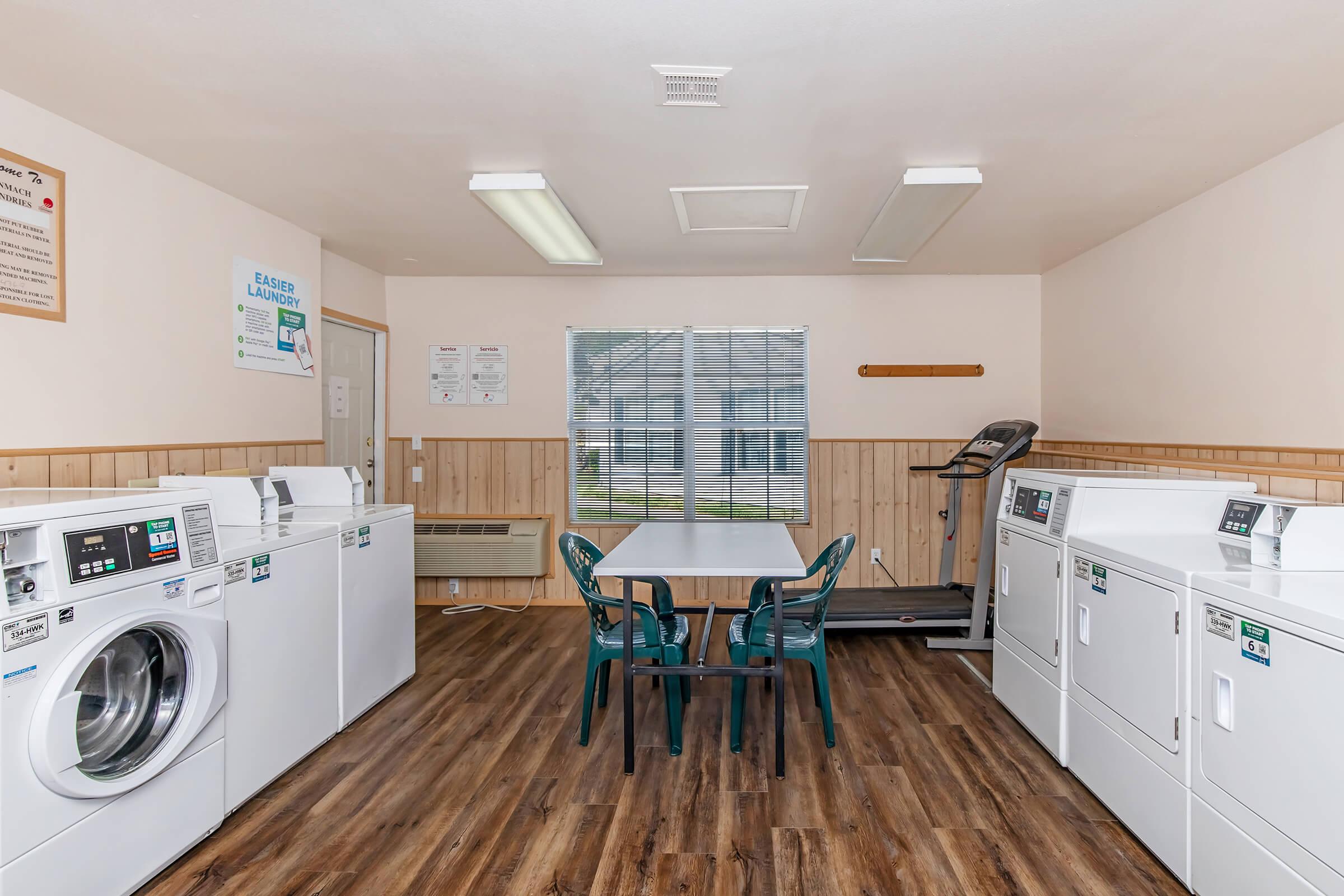 A clean laundry room featuring two coin-operated washing machines and two dryers. There's a table with two green chairs in the center and a window letting in natural light. A treadmill is placed in one corner, adding to the amenities of the space. Wood paneling accentuates the walls.