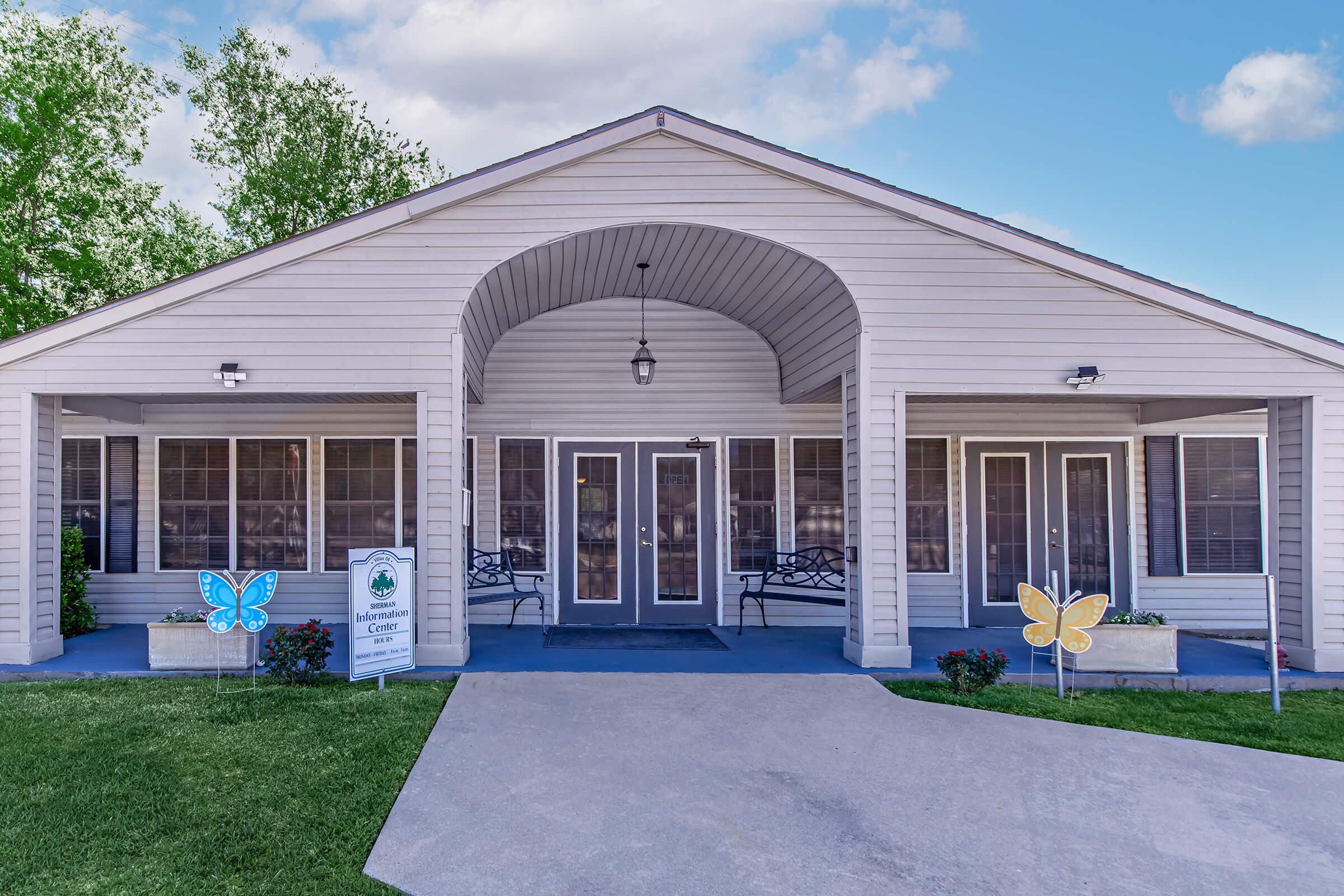 A light-colored, single-story building with a covered porch and multiple doors. The entrance features decorative benches and flower arrangements. Two large, colorful butterfly decorations are positioned on either side of the walkway, adding a cheerful touch. Lush greenery surrounds the building, and the sky is bright and clear.