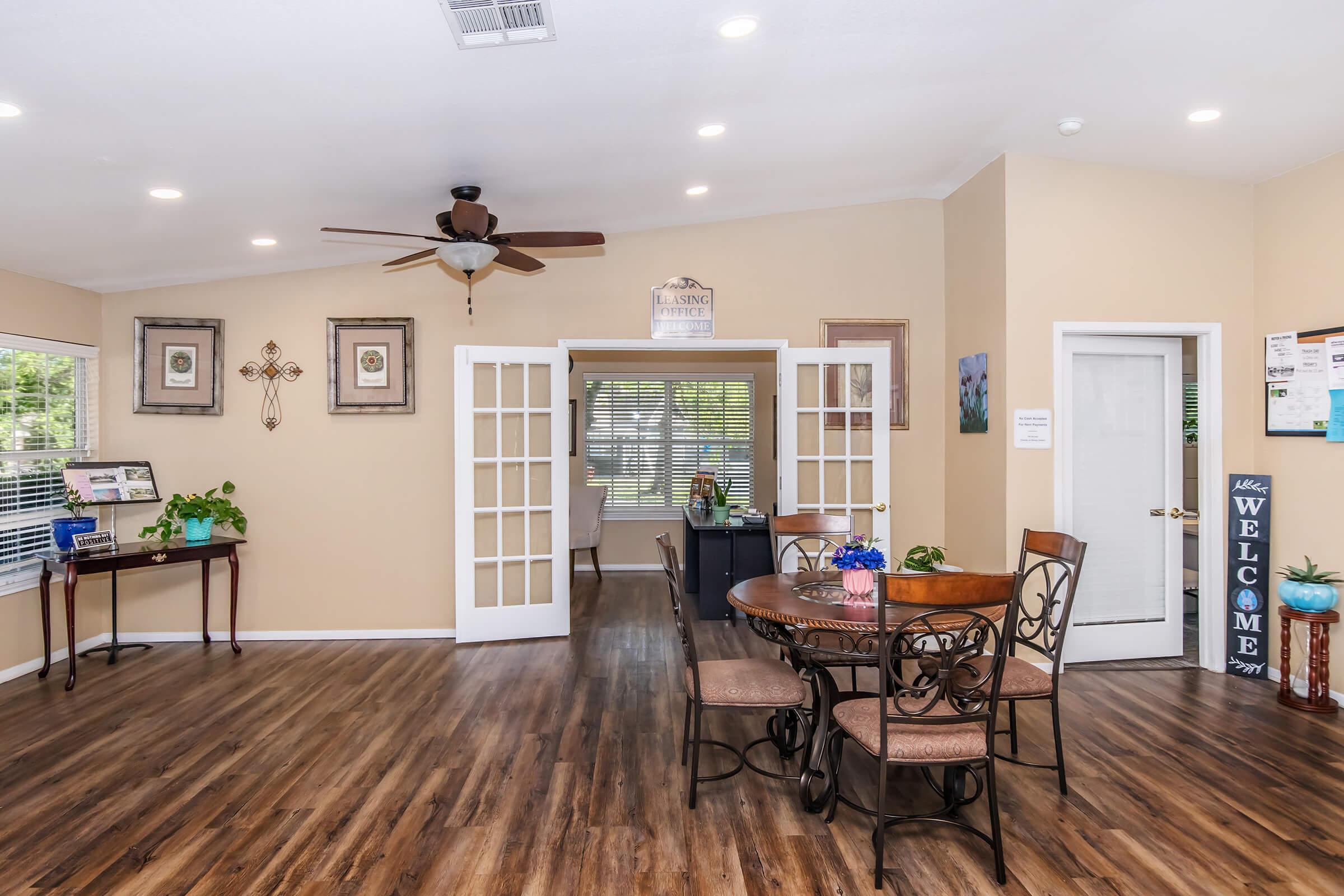 A spacious interior of a welcoming room with wooden flooring, featuring a round dining table with four chairs, plants, and decorative wall art. French doors lead to another area, and a sign displaying "Welcome" is visible. Soft lighting from the ceiling creates a warm atmosphere.