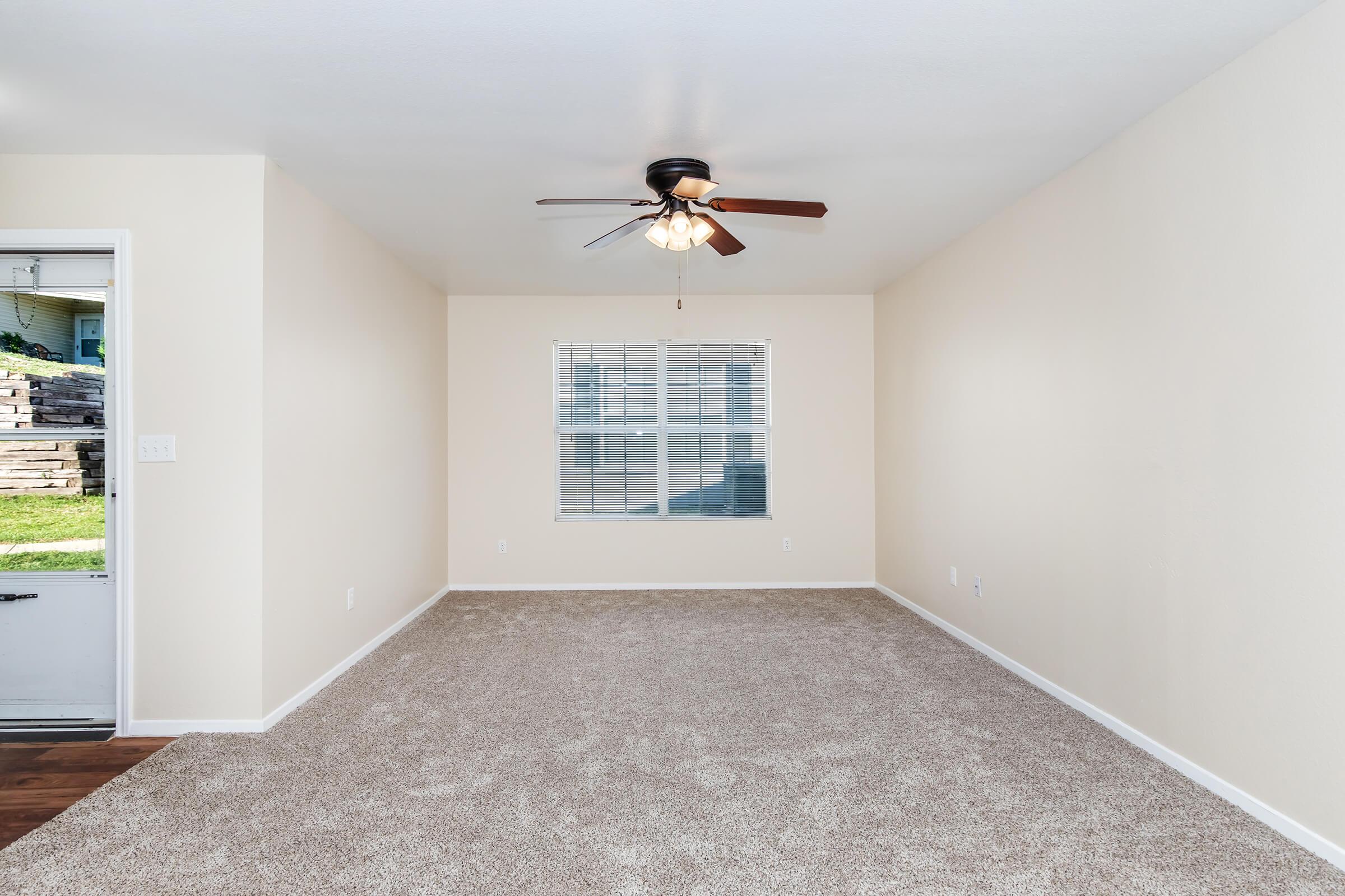 Empty living room with light beige walls and a ceiling fan. The space features tan carpet and a window with white blinds, allowing natural light. A door on the left leads outside, creating an open and airy atmosphere.