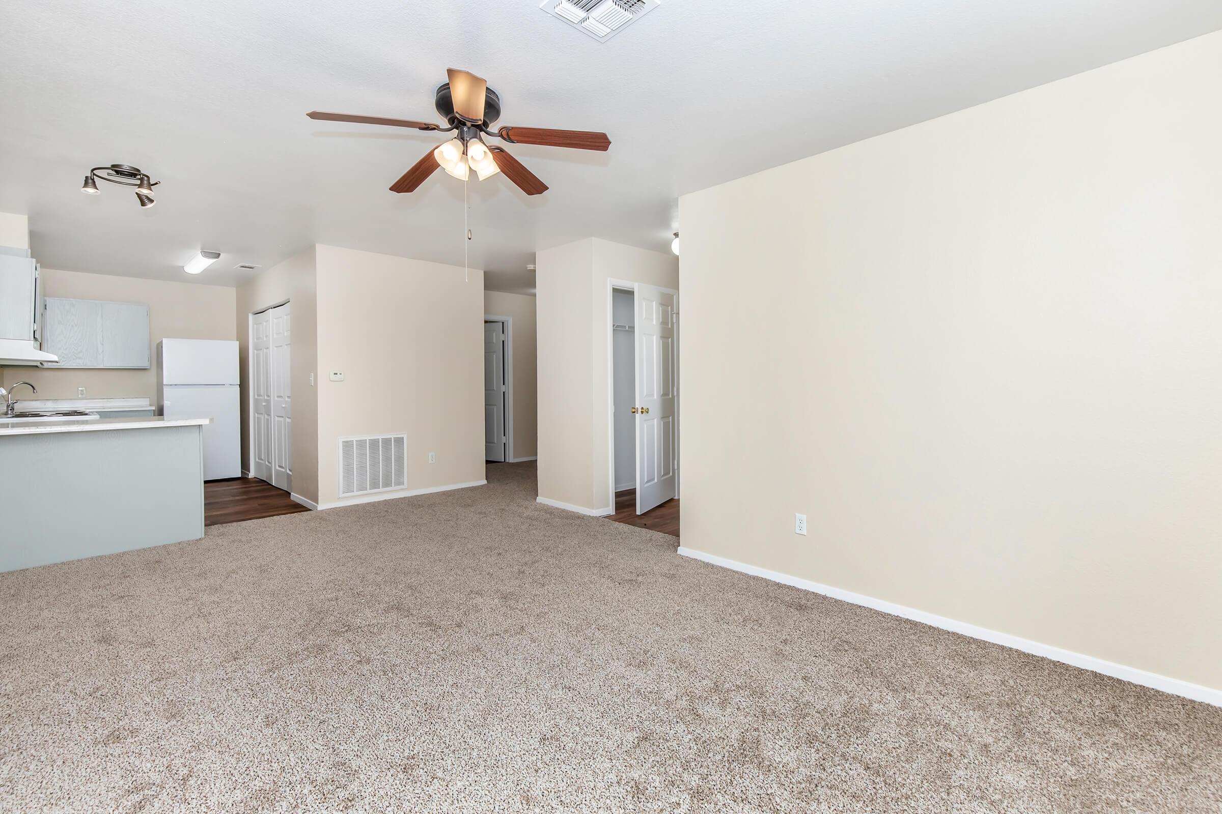 A spacious, well-lit living area featuring light beige walls and plush carpet. A ceiling fan hangs above, and the open layout connects to a kitchen with light cabinetry. Doorways lead to other rooms, and natural light enters through windows, creating a warm and inviting atmosphere.