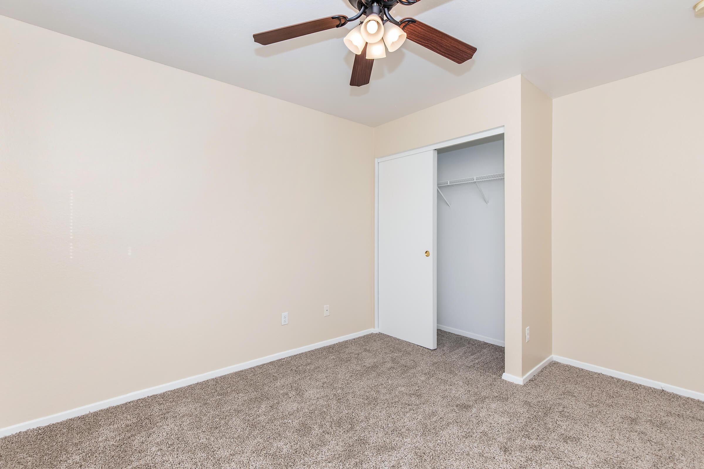 A spacious, empty room with beige walls and light brown carpet. The ceiling features a ceiling fan with light fixtures. There’s a white closet door on the left, and the room is well-lit with natural light.