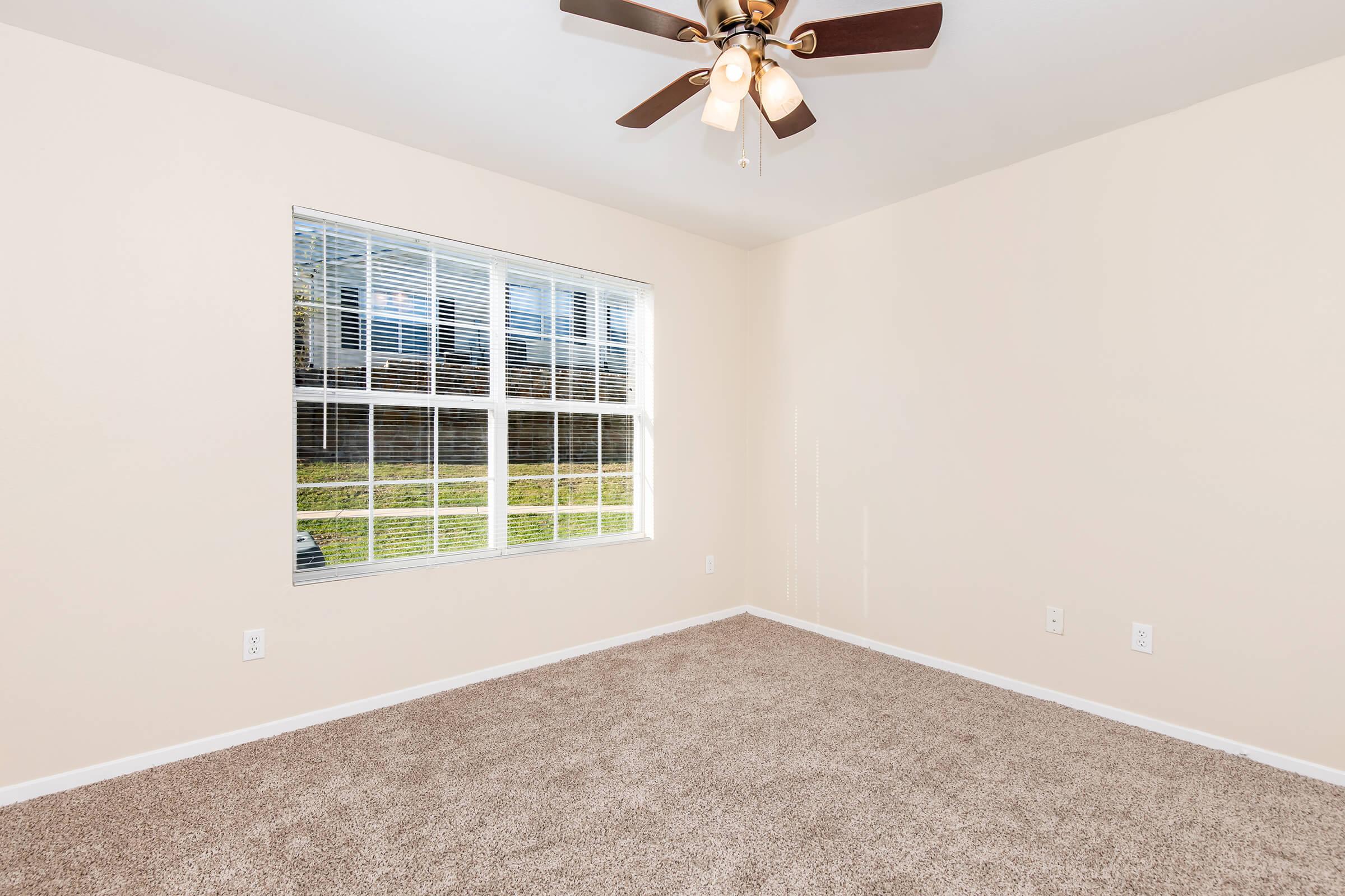 Empty room with light beige walls and a plush carpet. A large window with multiple panes lets in natural light, and a ceiling fan with light fixtures hangs from the ceiling. The room is uncluttered and ready for furniture placement. Outdoor greenery is visible through the window.