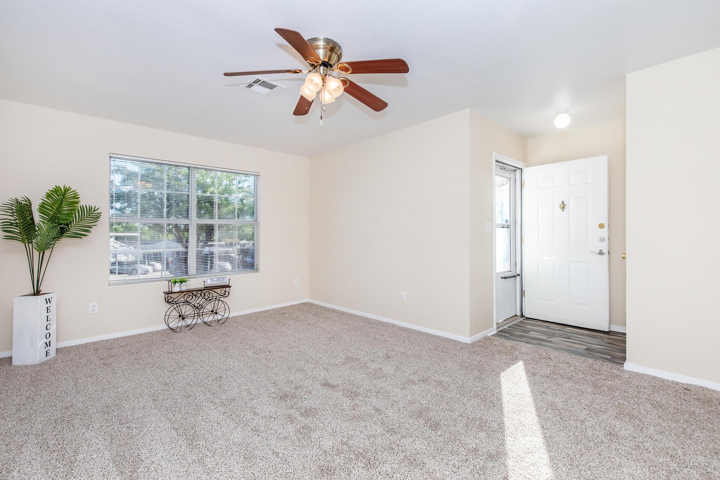A bright, inviting room with beige walls and plush carpet. A ceiling fan with light hangs above. There's a window allowing natural light in, a small table with a plant beside it, and a front door with a glass panel. The overall atmosphere is warm and welcoming, ideal for a cozy living space.