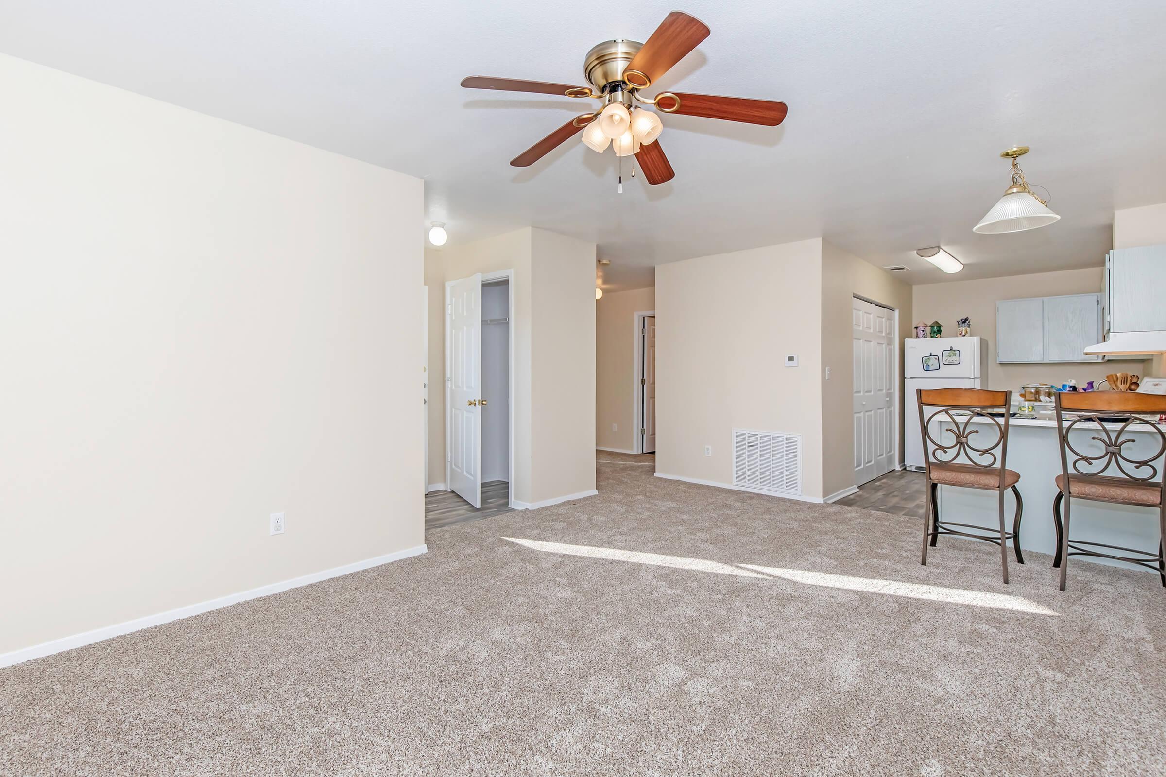Spacious living area featuring a ceiling fan, light-colored walls, and plush carpeting. A small kitchen with bar stools is visible in the background, along with doors leading to other rooms. Bright lighting enhances the open feel of the space.