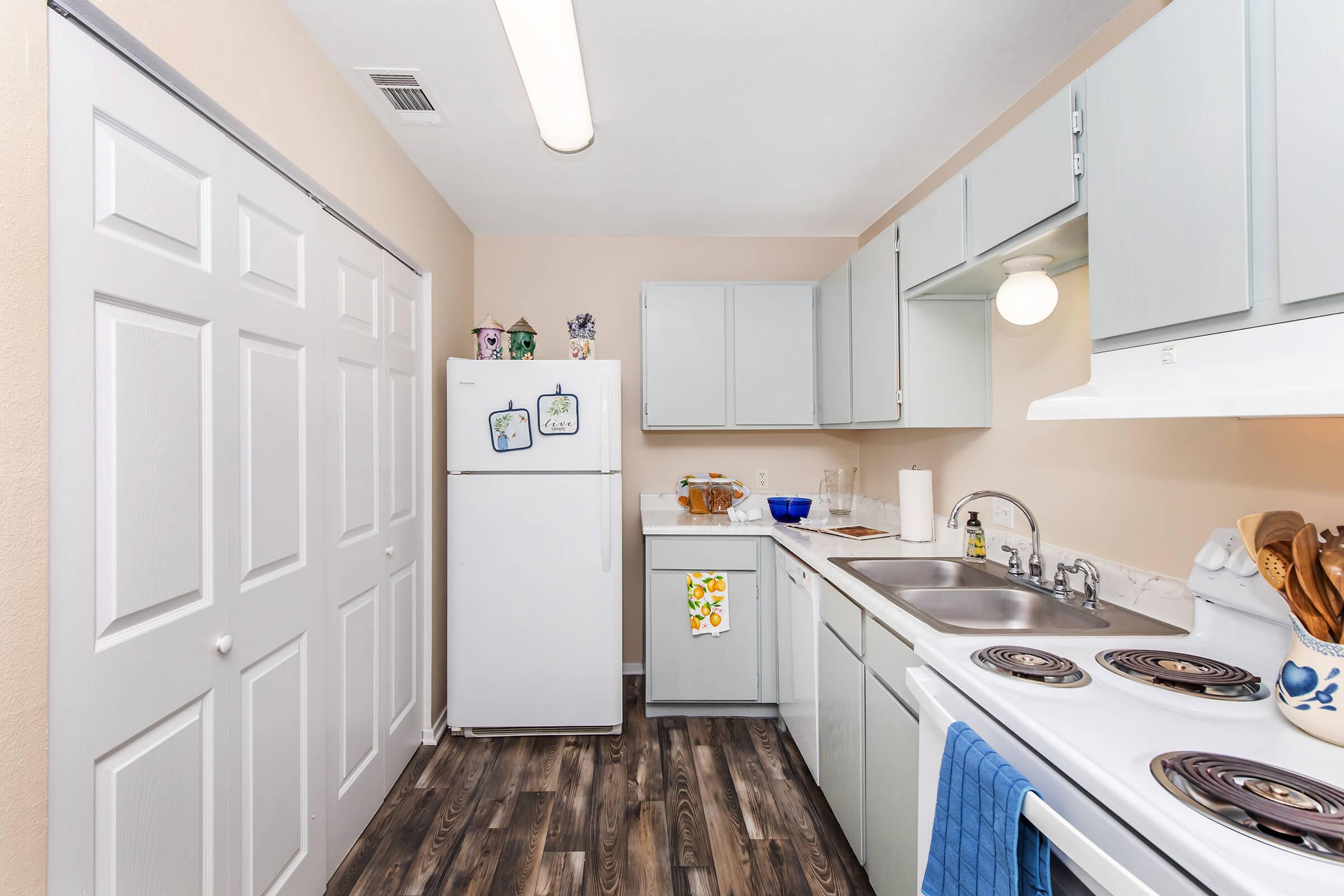 A modern kitchen featuring light gray cabinets, a white refrigerator, and a white stove with an oven. The countertops are clean and organized, displaying utensils. The floor has a wood-like appearance, and there are decorative items and a dish towel hanging by the sink. The walls are painted a soft beige.