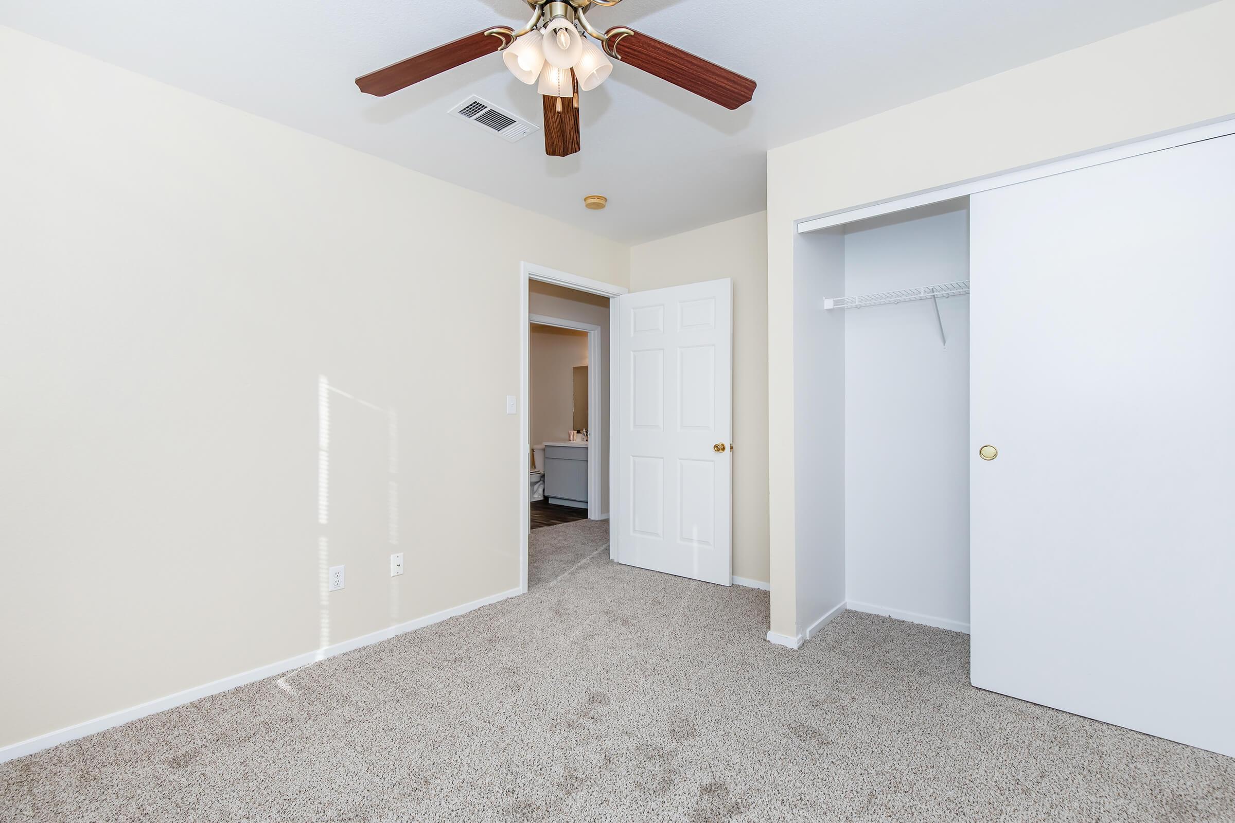 A well-lit bedroom featuring beige walls, carpeted flooring, and a ceiling fan with wooden blades. The room includes a door leading to another area, and a closet with sliding doors. Natural light enters through a window, creating a warm atmosphere.