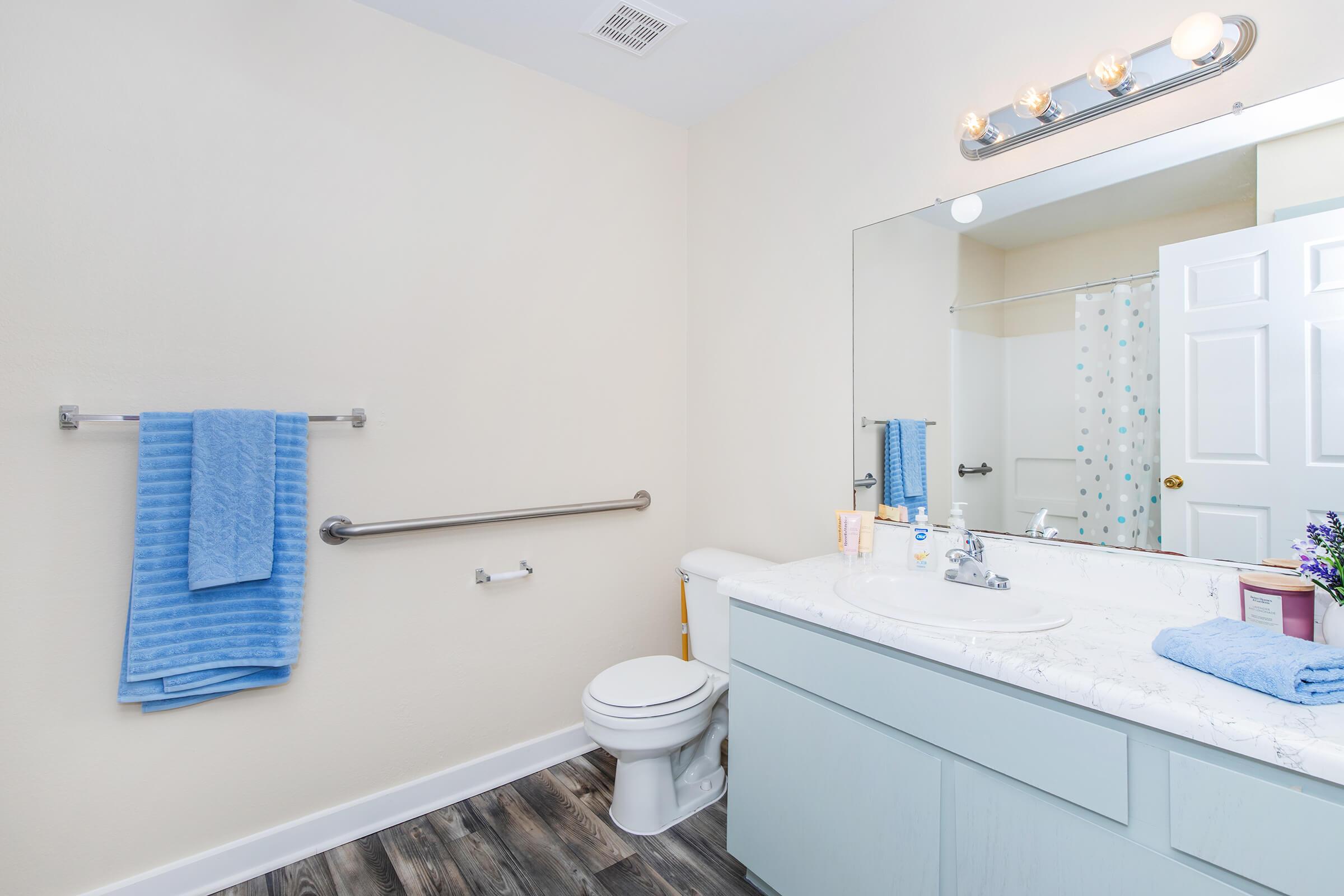 A clean, modern bathroom featuring a light-colored wall, a large mirror above the sink, a white toilet, and a blue towel hanging on a rack. The sink has a marble countertop with toiletries and a small vase of flowers, while the floor is covered with dark wood-like tiles. A shower curtain with a colorful polka dot pattern is visible.