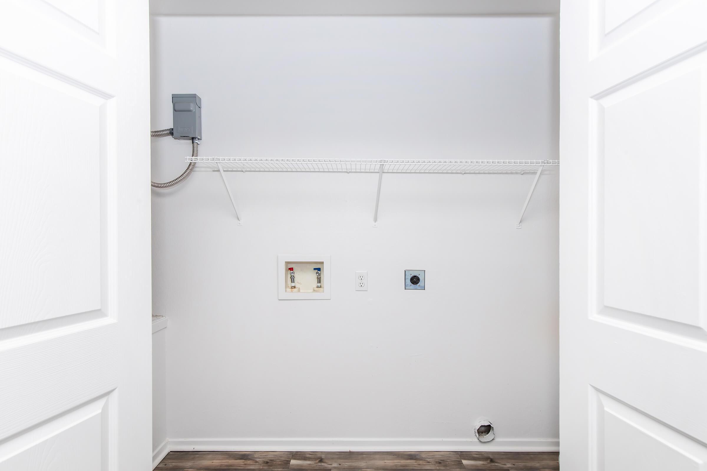A clean, empty laundry room with white walls and a light wooden floor. Two open closet doors reveal a shelf supported by metal brackets, an electrical panel on the left, and two wall outlets. There is a vent on the floor, and the space is well-lit and organized.