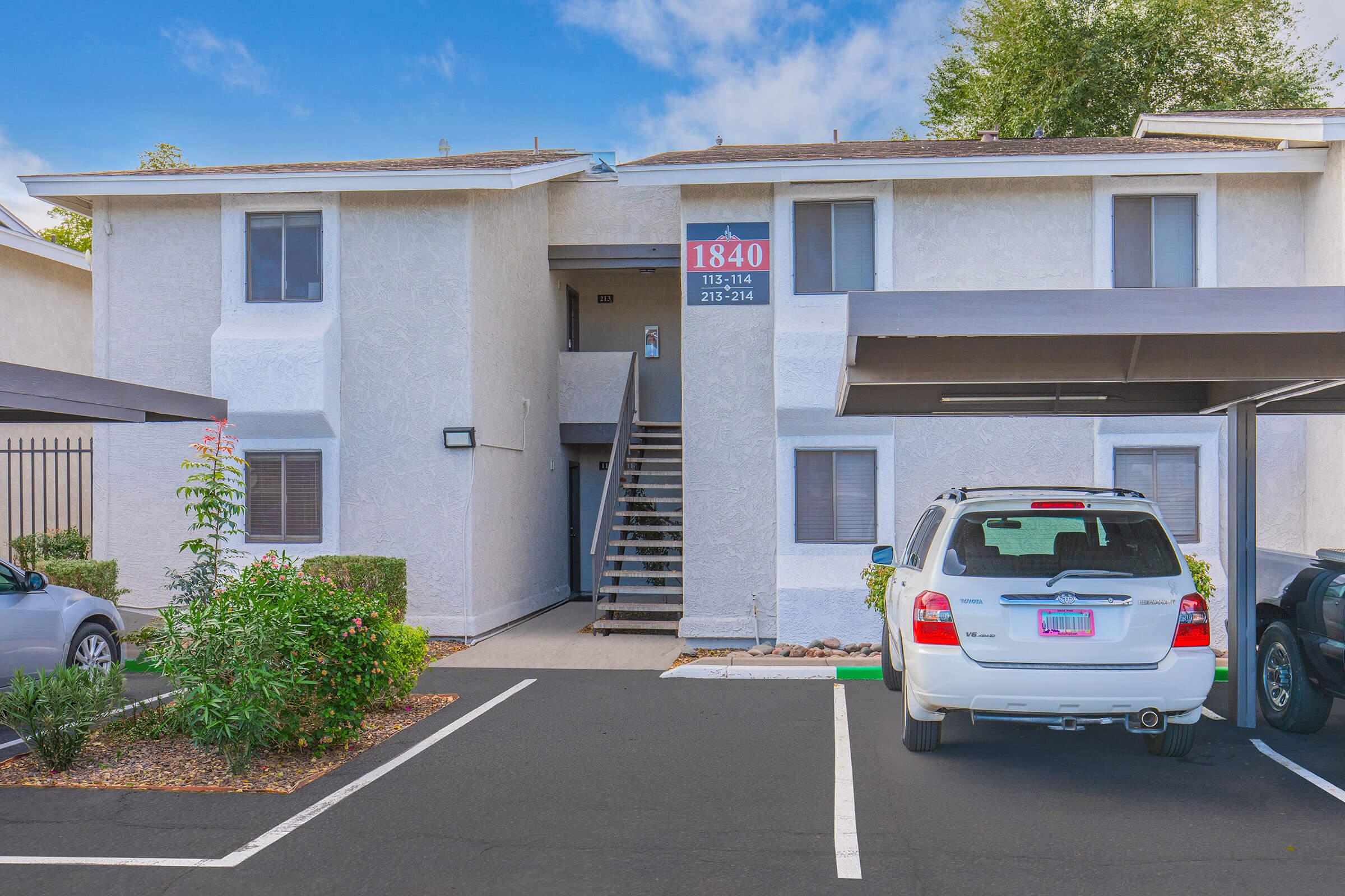 A multi-unit residential building with a light-colored exterior. The building features a central staircase leading to upper units, and a parking area with several vehicles, including a white SUV and a grey car. The address "1840" is prominently displayed on the building, along with contact information.