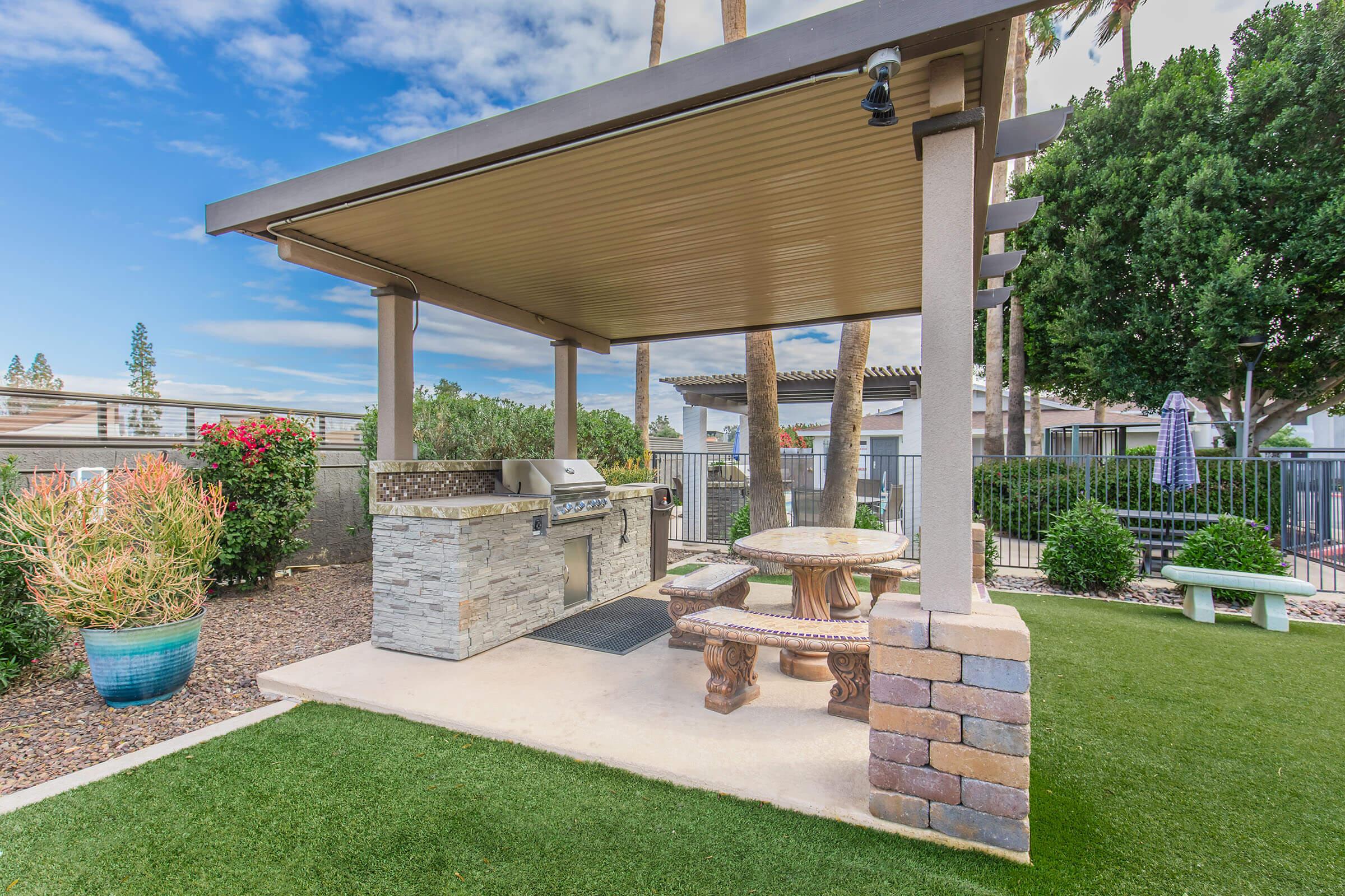 A landscaped outdoor area featuring a covered barbecue grill station with a stone countertop and surrounding seating. There are decorative plants, a green lawn, and a clear blue sky in the background, creating a pleasant and inviting atmosphere for outdoor dining and gatherings.