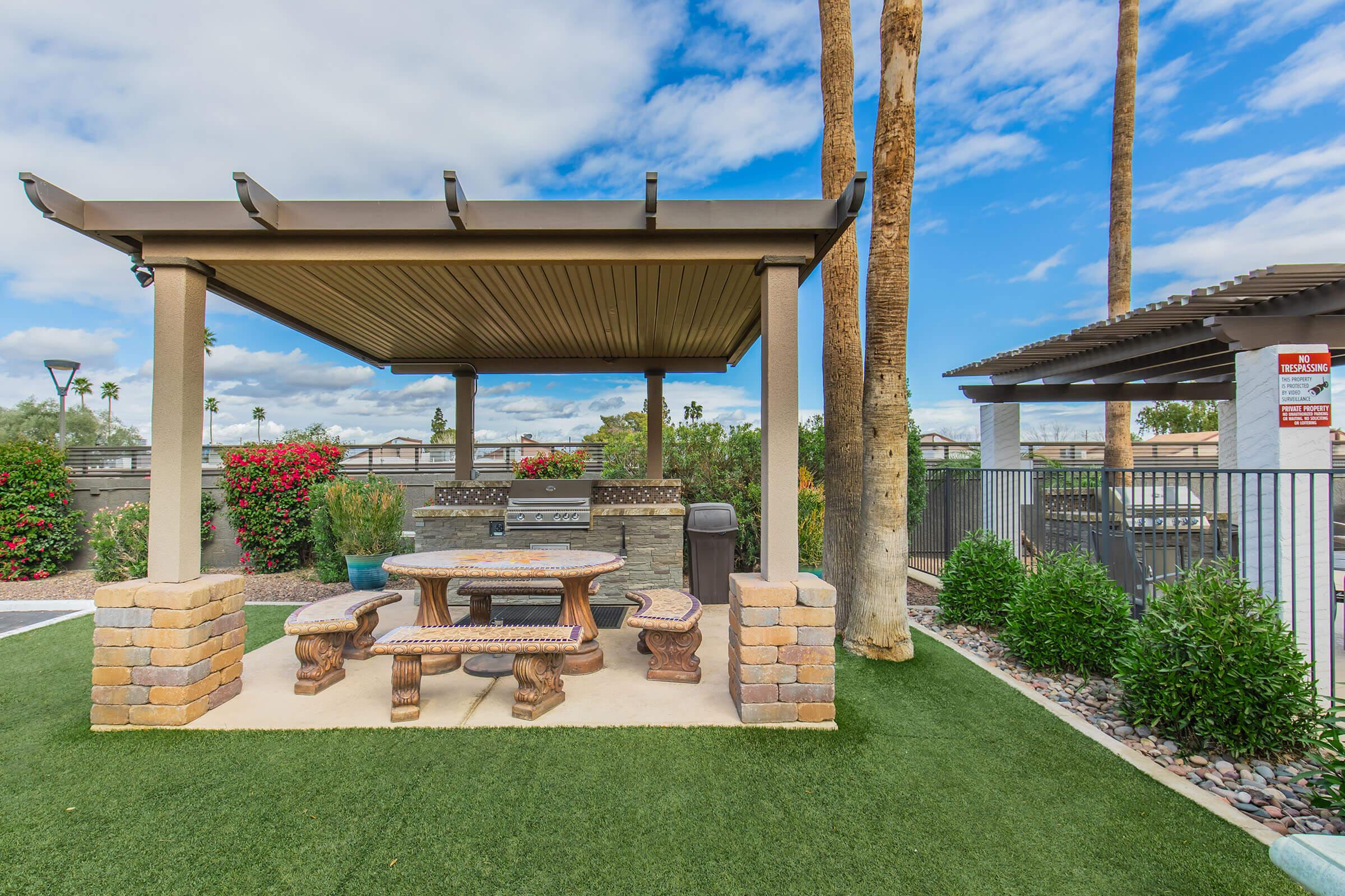 A shaded outdoor seating area featuring a stone table and benches surrounded by lush greenery. In the background, there are palm trees and a clear blue sky, along with a fence and flowering plants adding color to the scene. Perfect for relaxation or gatherings.