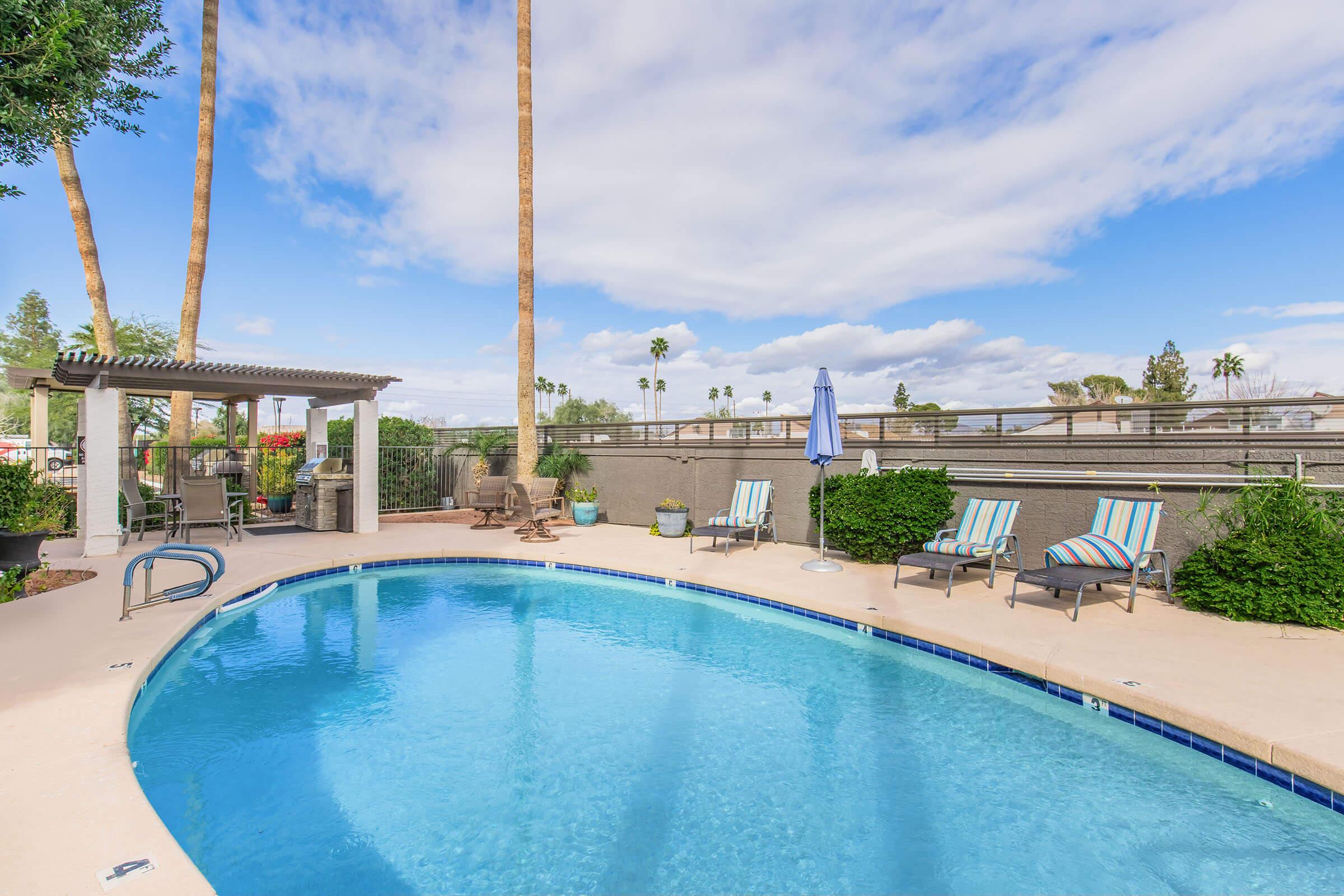 A sparkling blue swimming pool surrounded by palm trees and lounge chairs. A shaded seating area with a pergola is visible in the background, along with decorative plants and a clear blue sky. The scene conveys a relaxing outdoor atmosphere perfect for leisure.