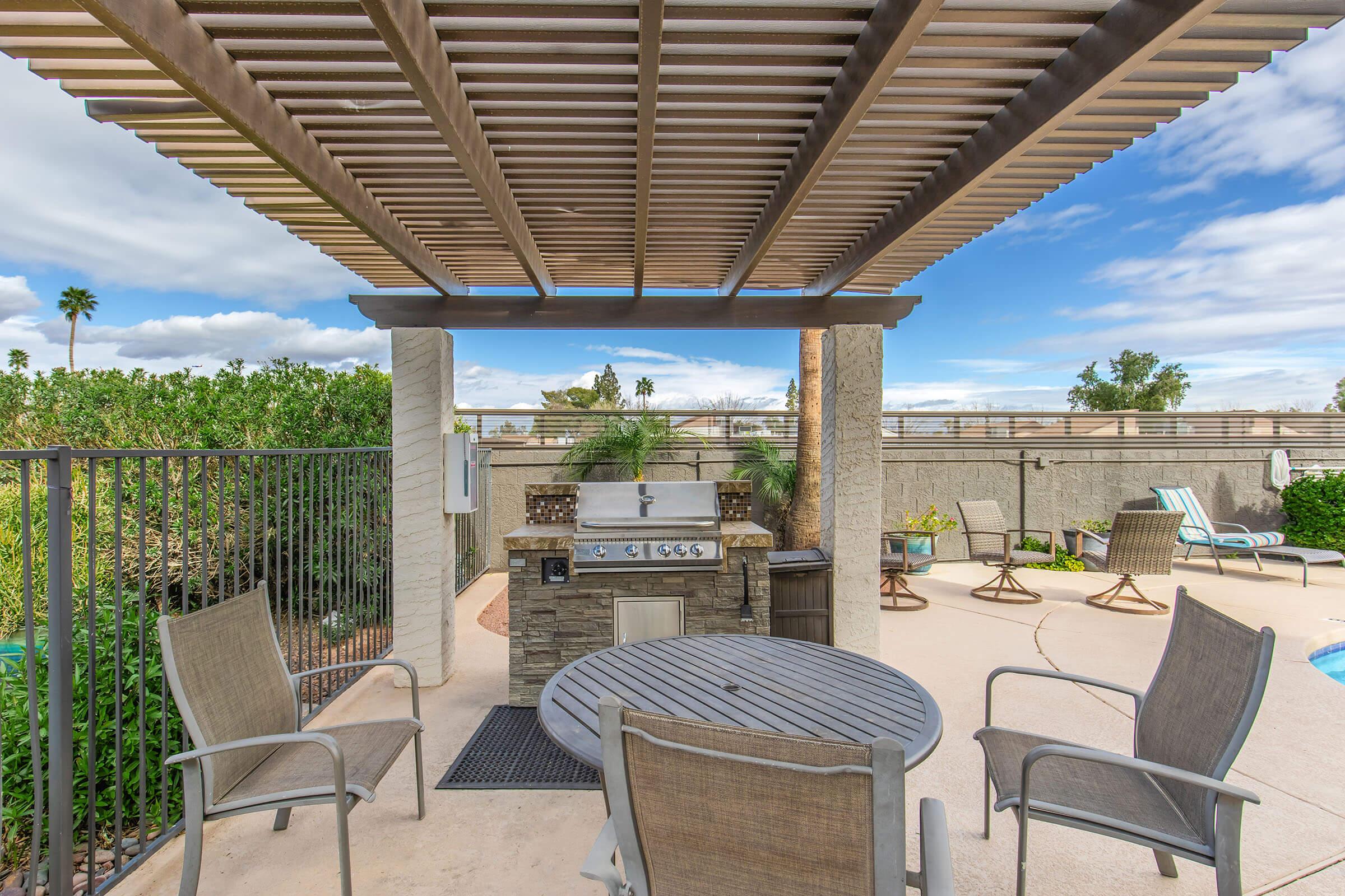 Outdoor patio area featuring a stone barbecue grill under a pergola. Surrounding the grill are four chairs and a round wooden table. The scene includes green foliage and palm trees in the background, along with a clear blue sky and wispy clouds, creating a relaxing outdoor setting.