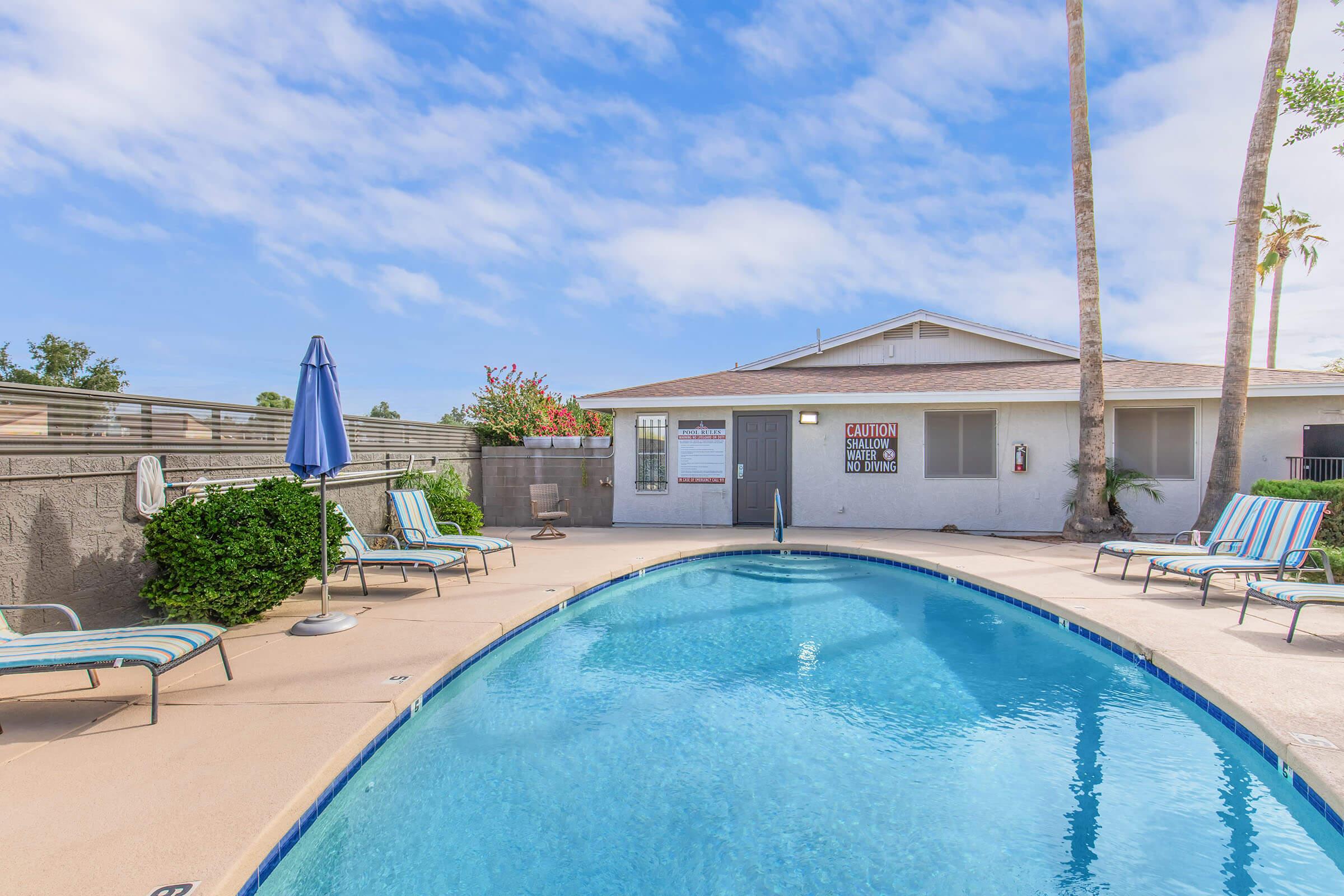 A well-maintained swimming pool surrounded by lounge chairs and a sun umbrella. In the background, there is a building with a caution sign and lush greenery. Palm trees line the area under a clear blue sky.