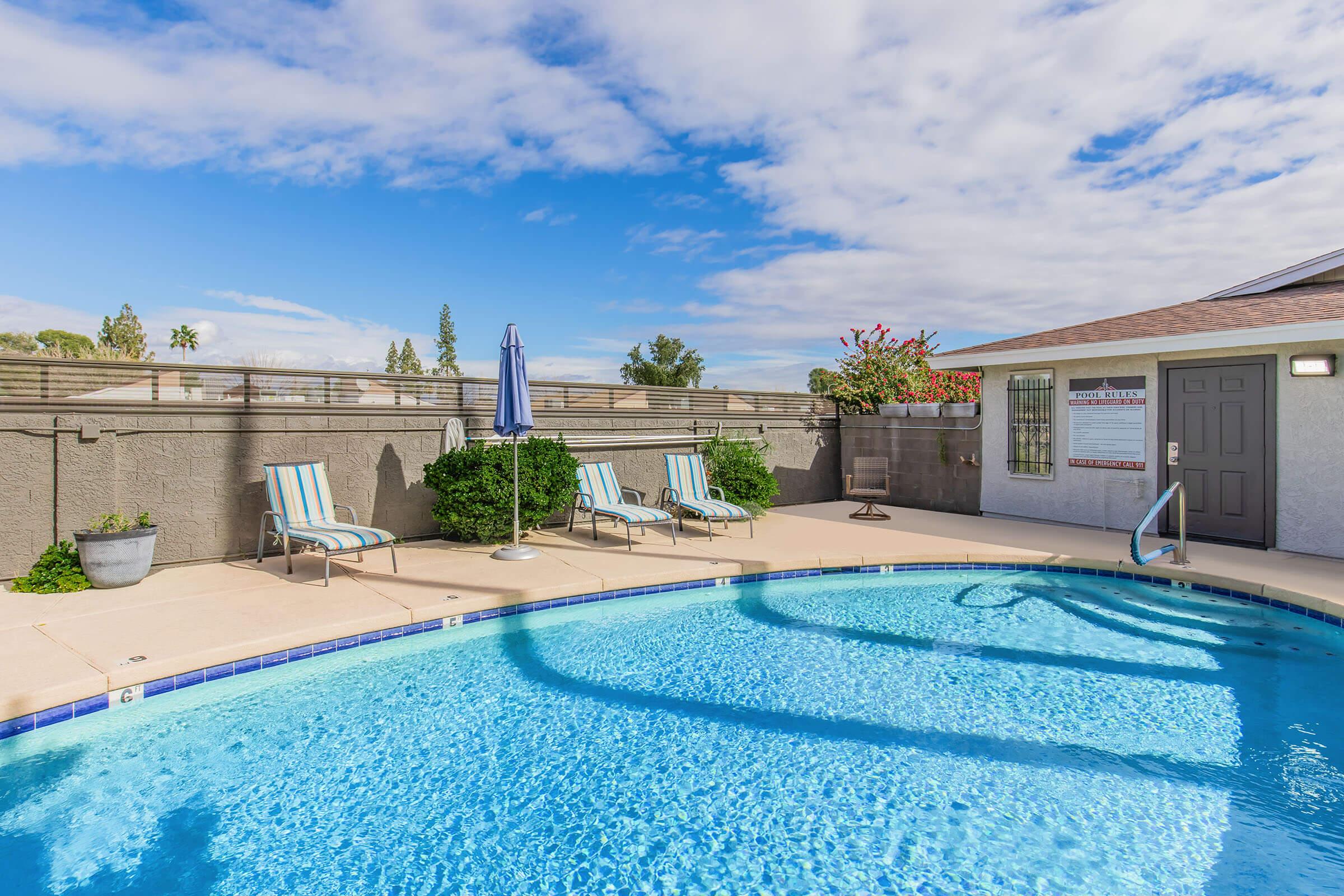 A serene outdoor pool area featuring clear blue water, lounge chairs with striped fabric, and a sun umbrella. The surrounding patio is well-maintained, with a wall of greenery and a building with a door and a sign in the background under a partly cloudy sky.