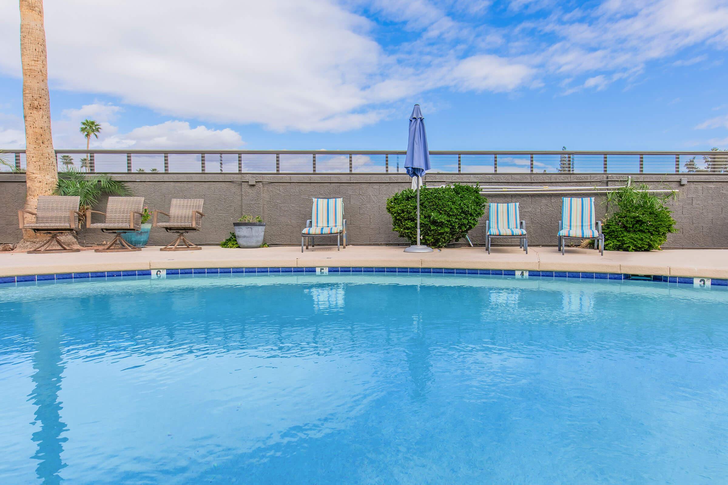 A clear blue swimming pool with lounge chairs arranged nearby. An umbrella stands by the poolside, and there are trees and plants in the background against a partly cloudy sky. The overall scene conveys a relaxing outdoor setting.