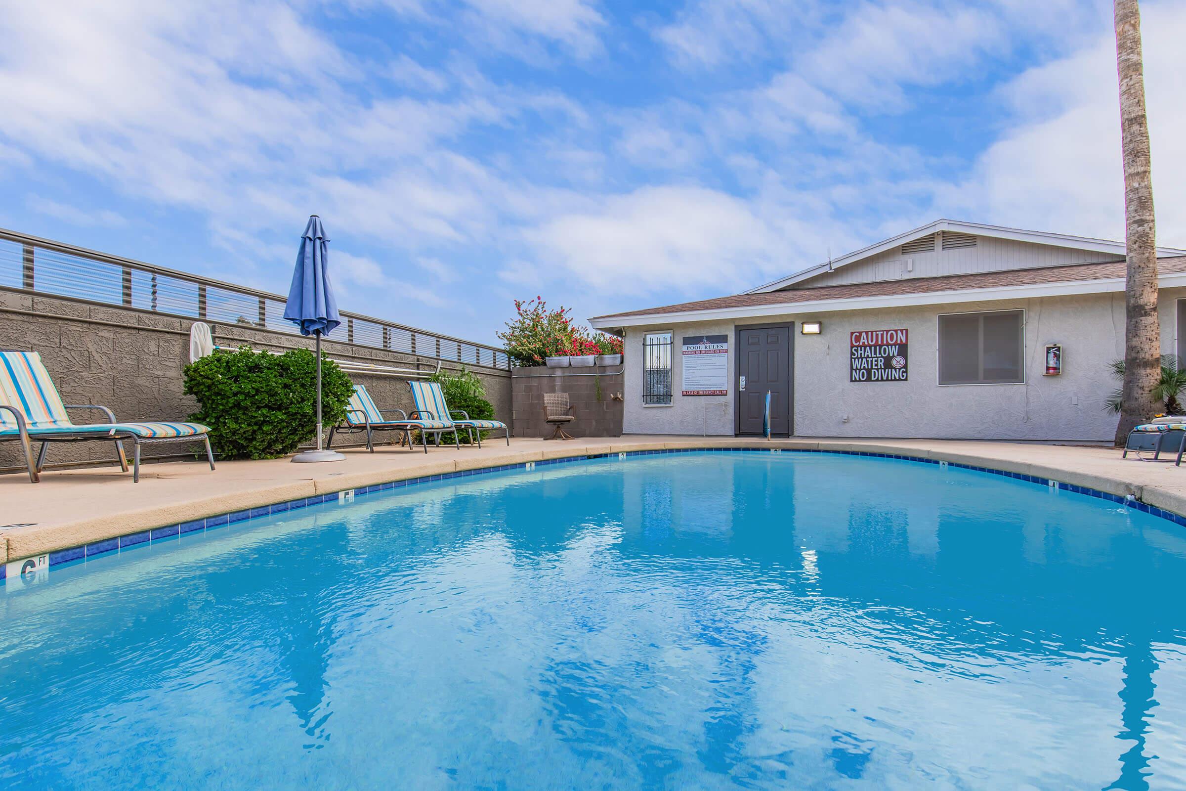 A clear blue swimming pool surrounded by lounge chairs, with a building in the background displaying a caution sign. There are palm trees and a gray sky, creating a relaxing atmosphere. The area appears well-maintained and inviting for leisure activities.