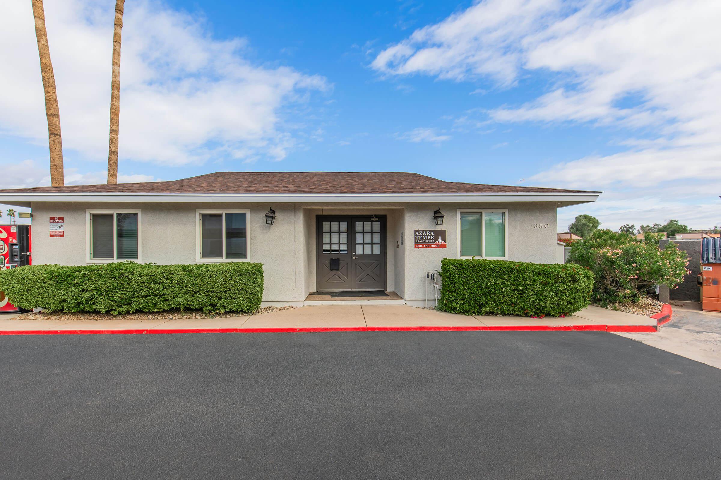 A single-story building with a gray exterior, featuring a dark brown roof and two windows. There are neatly trimmed green shrubs in front of the building, and a parking lot with a red curb. Palm trees are visible in the background under a blue sky with scattered clouds.