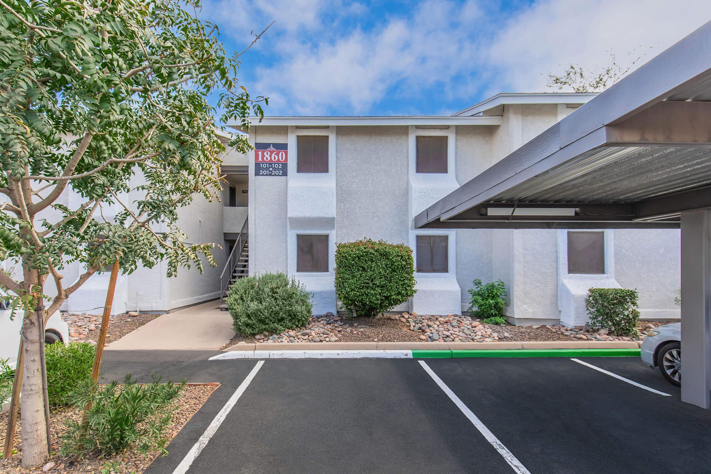 Exterior view of a two-story apartment building with a for-rent sign displaying the number "1860." The building features white walls, several windows, and a covered parking area. Lush greenery surrounds the entrance and parking lot, and the sky is partly cloudy.